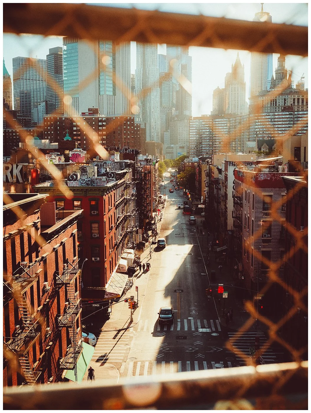 Lower Manhattan seen through the weathered fencing of Manhattan Bridge, where late-afternoon light turns the city into layers of steel, brick and shadow.