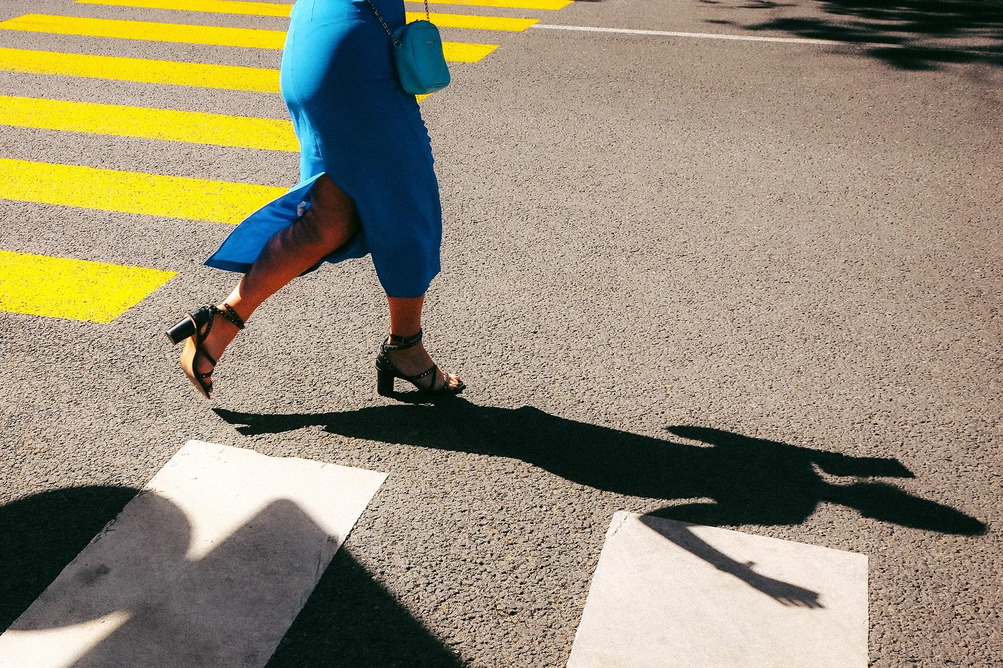 Woman in a blue dress crossing a sunlit street, with yellow road markings and a long shadow.