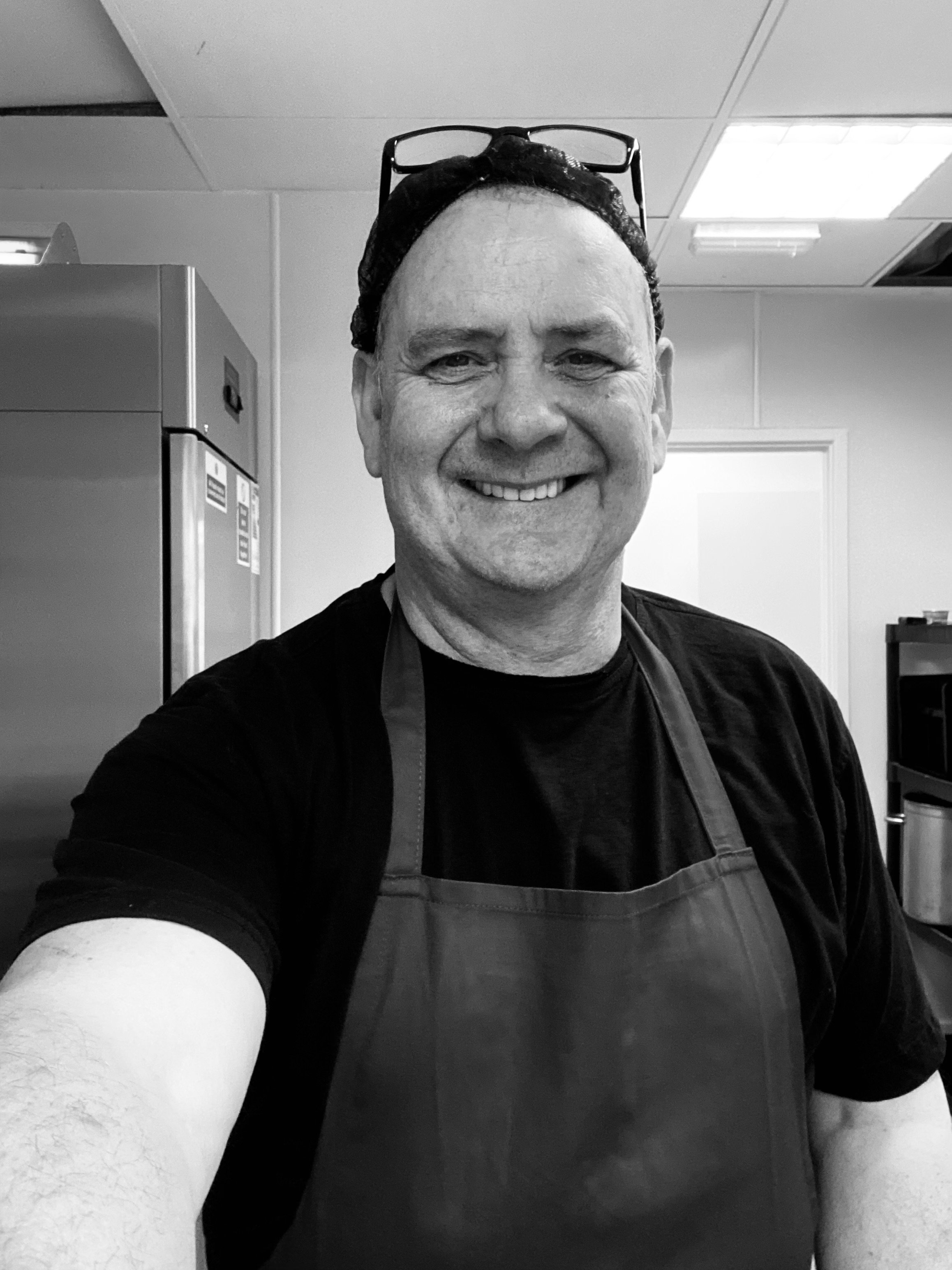 Black and white photo of a smiling man wearing glasses on his head, a black shirt, and a striped apron, in a kitchen.