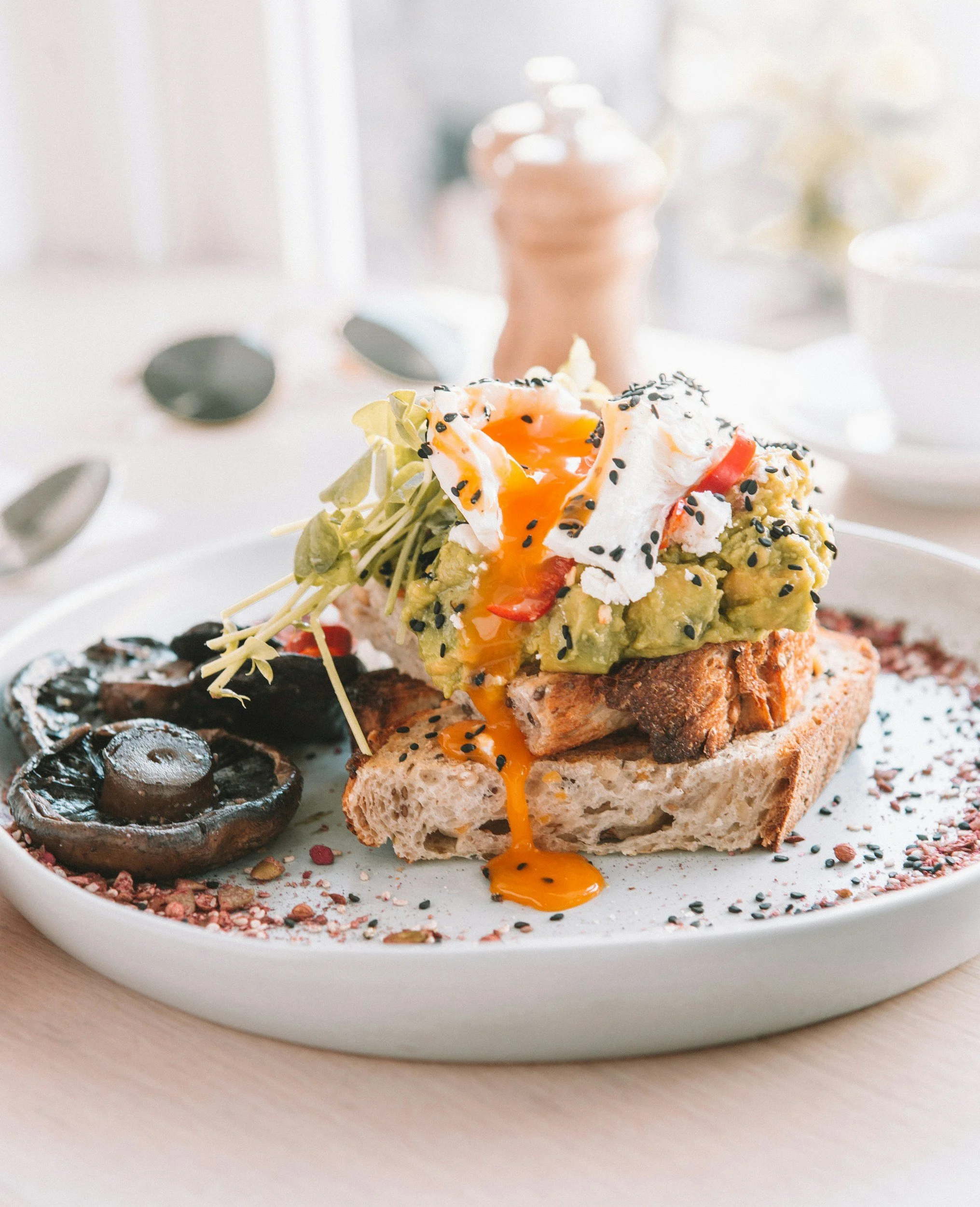 Close-up of a slice of avocado toast topped with a poached egg, red pepper slices, and black sesame seeds on a white plate. The yolk is slightly runny and spilling over the bread. In the background, there are blurred glass salt and pepper shakers.