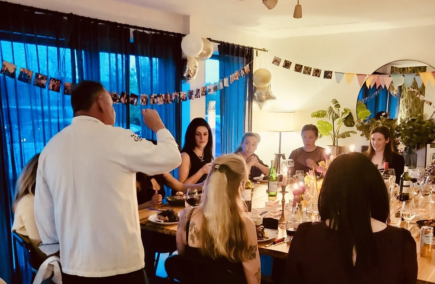 People gathered around a dinner table during a celebration, decorated with balloons, photos, and colorful bunting.