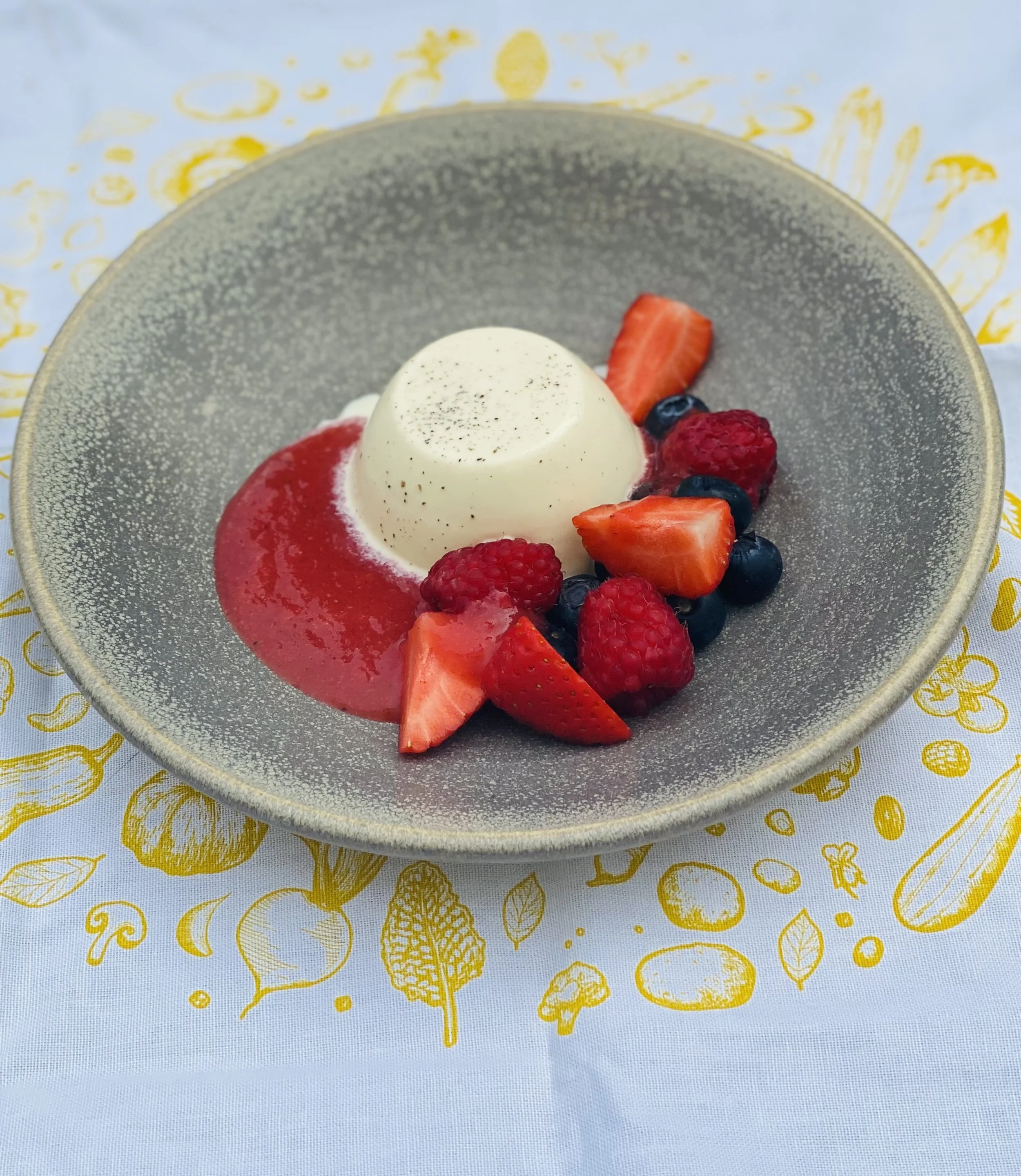 A bowl of panna cotta topped with mixed berries and strawberry slices, garnished with black pepper, on a white tablecloth with gold leaf patterns.