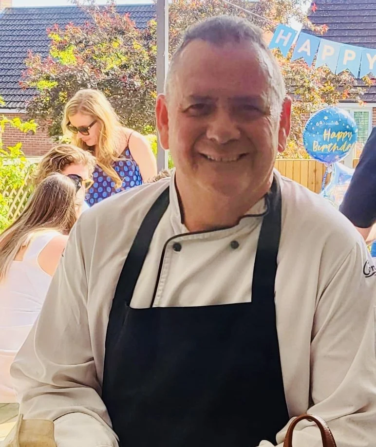 A chef in a white coat and black apron smiling during an outdoor birthday celebration. In the background, there are people sitting at a table, birthday balloons, and a banner that says 'HAPPY' in blue letters.
