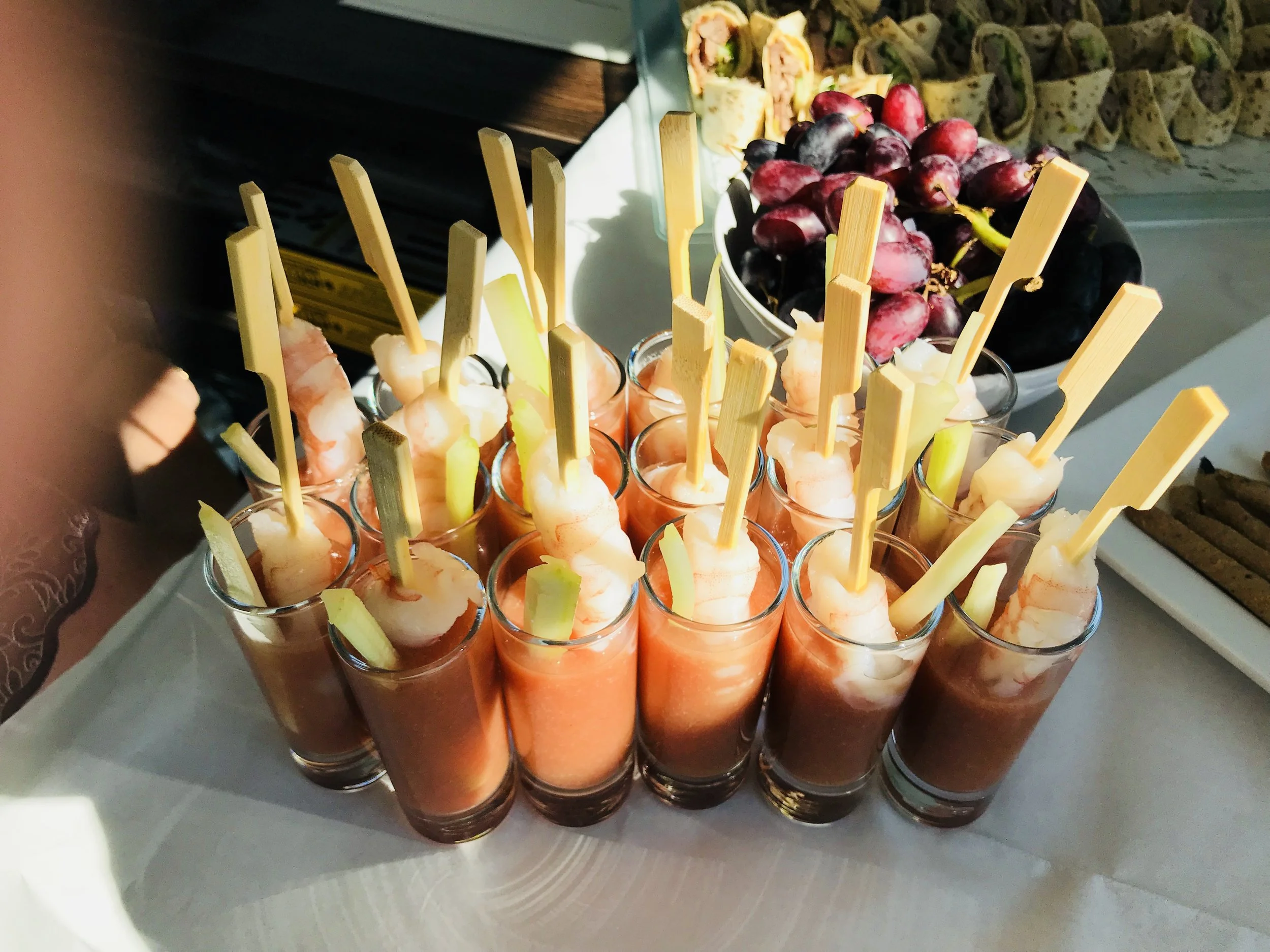 A tray of shrimp cocktails in small glasses with slices of pineapple and celery sticks, with a bowl of red and black grapes and a platter of wraps in the background.