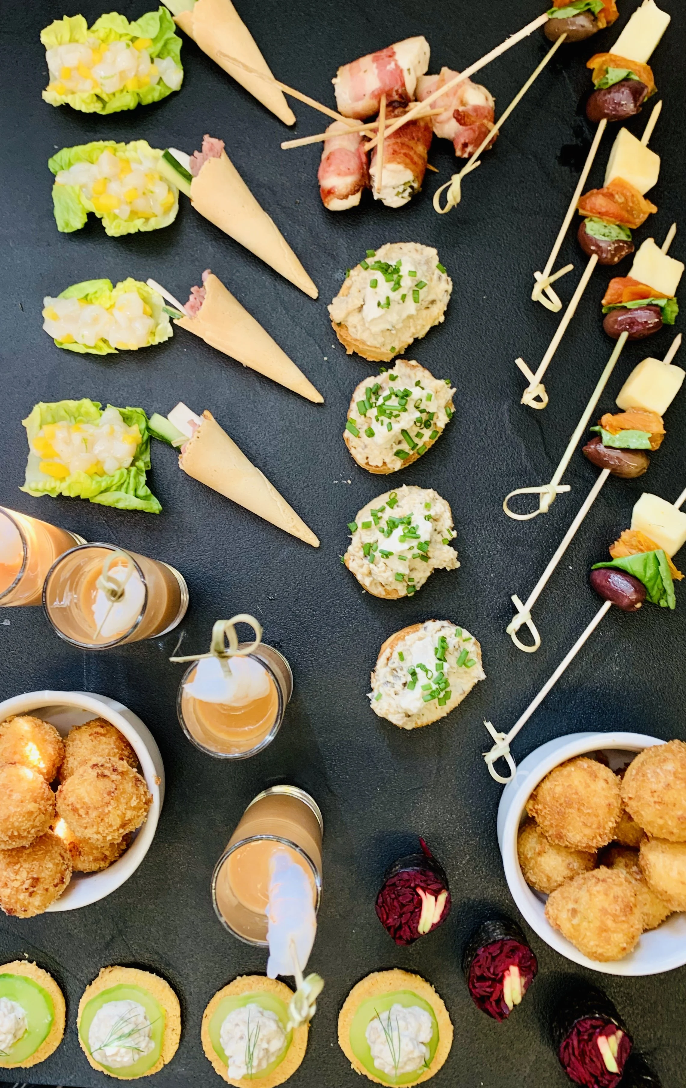 Assorted appetizers including small sandwiches with lettuce and spreads, rolled meat and cheese skewers, stuffed cherry tomatoes, and breaded fried snacks on a black serving tray.