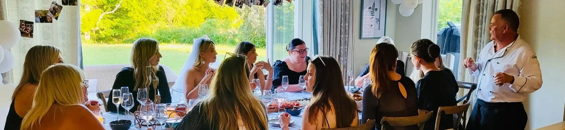 Group of women attending a celebration or party, sitting at a dining table with food and drinks, with a chef or host standing and talking to them, in a room with large windows showing a green outdoor area.