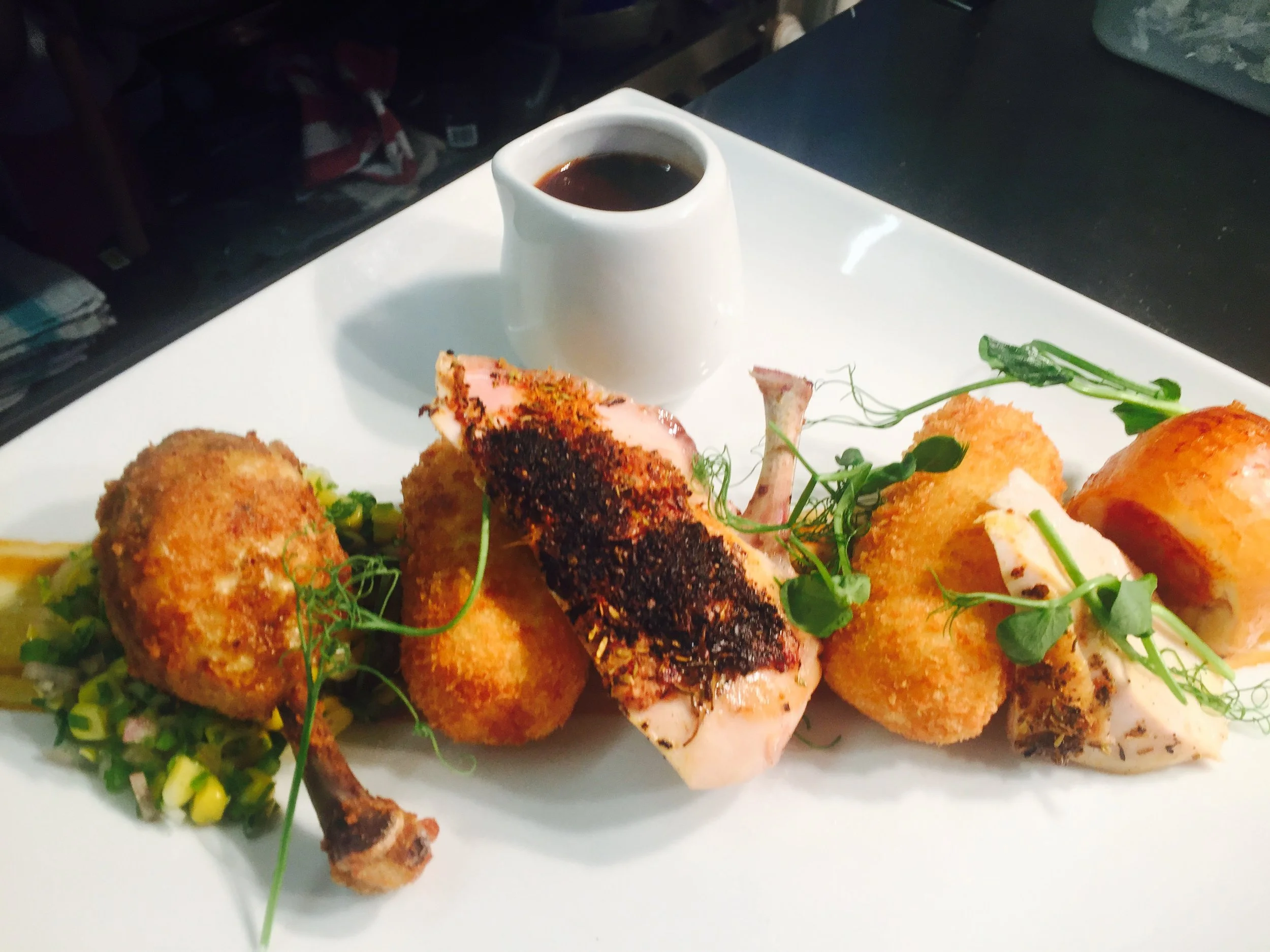 A white plate with a variety of breaded fried foods topped with microgreens, along with a small white creamer filled with dark sauce, on a table.