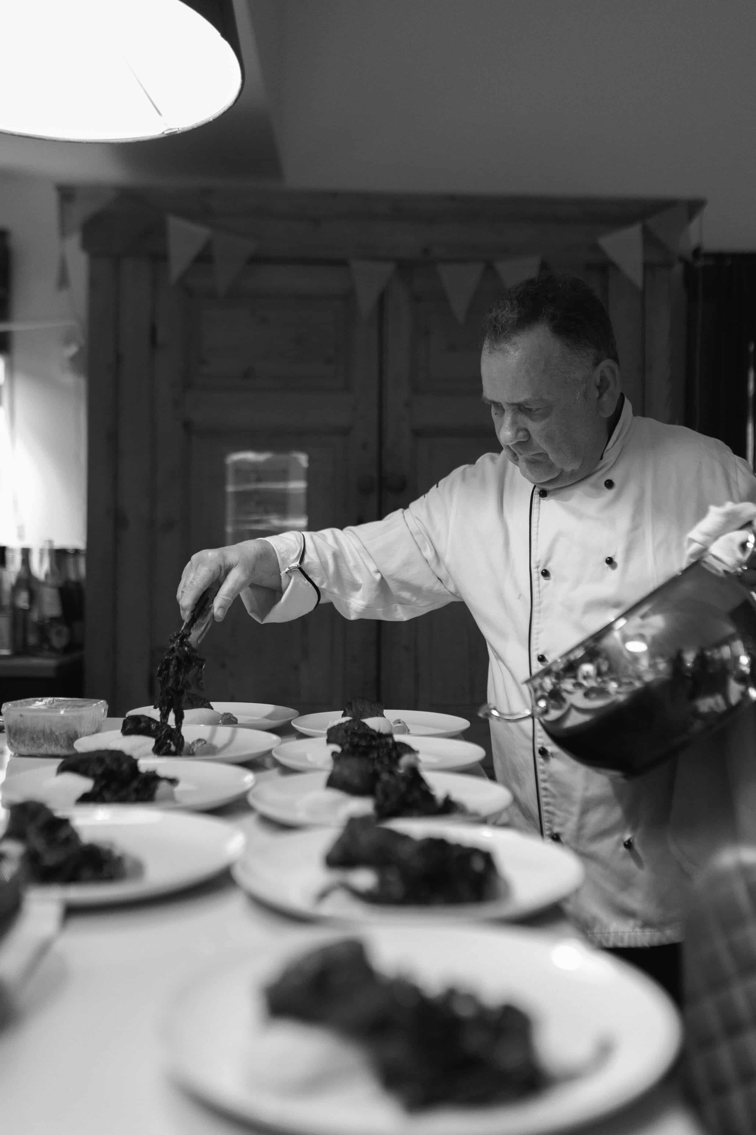 A professional chef in a white uniform garnished with herbs, preparing desserts or appetizers on a table with multiple small plates, surrounded by wooden cabinets and bunting decorations in a cozy kitchen or dining area.