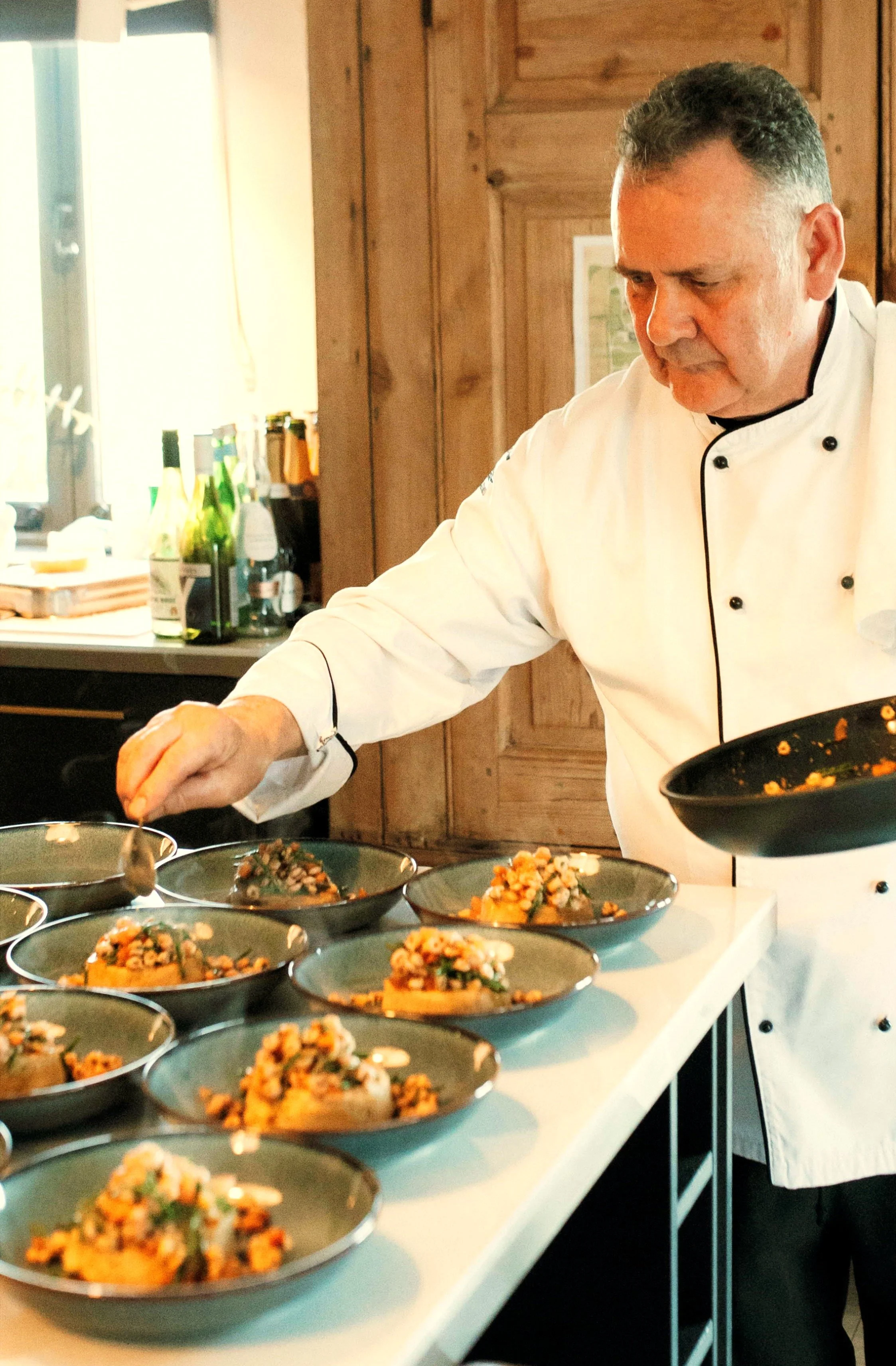 A chef in a white uniform preparing dishes in a restaurant kitchen with wooden walls, multiple plates of food on a white counter, and bottles on a shelf in the background.