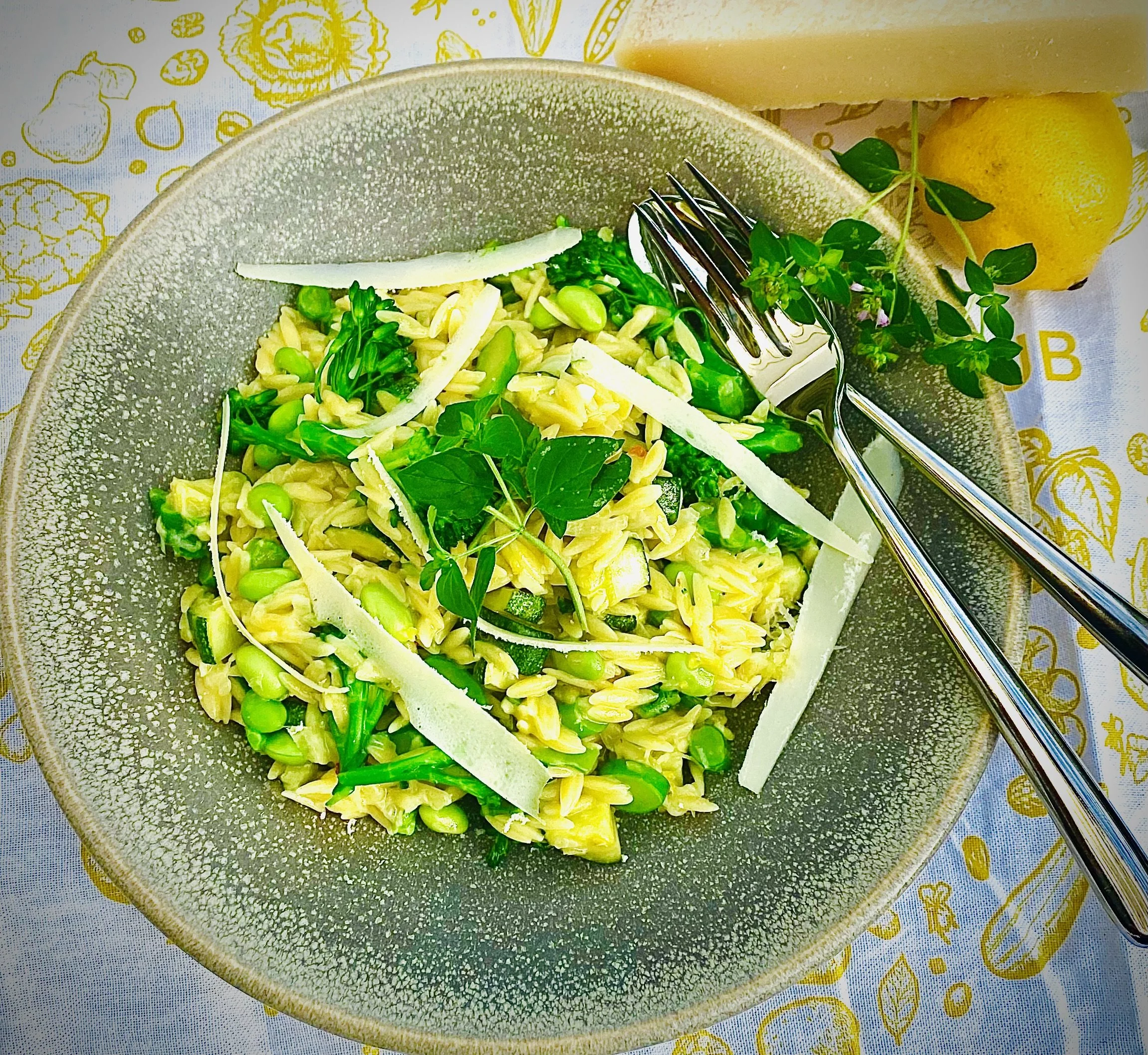 A bowl of pasta salad with green peas, shredded cheese, and herbs, on a yellow patterned tablecloth with lemon slices and a wedge of lemon nearby.