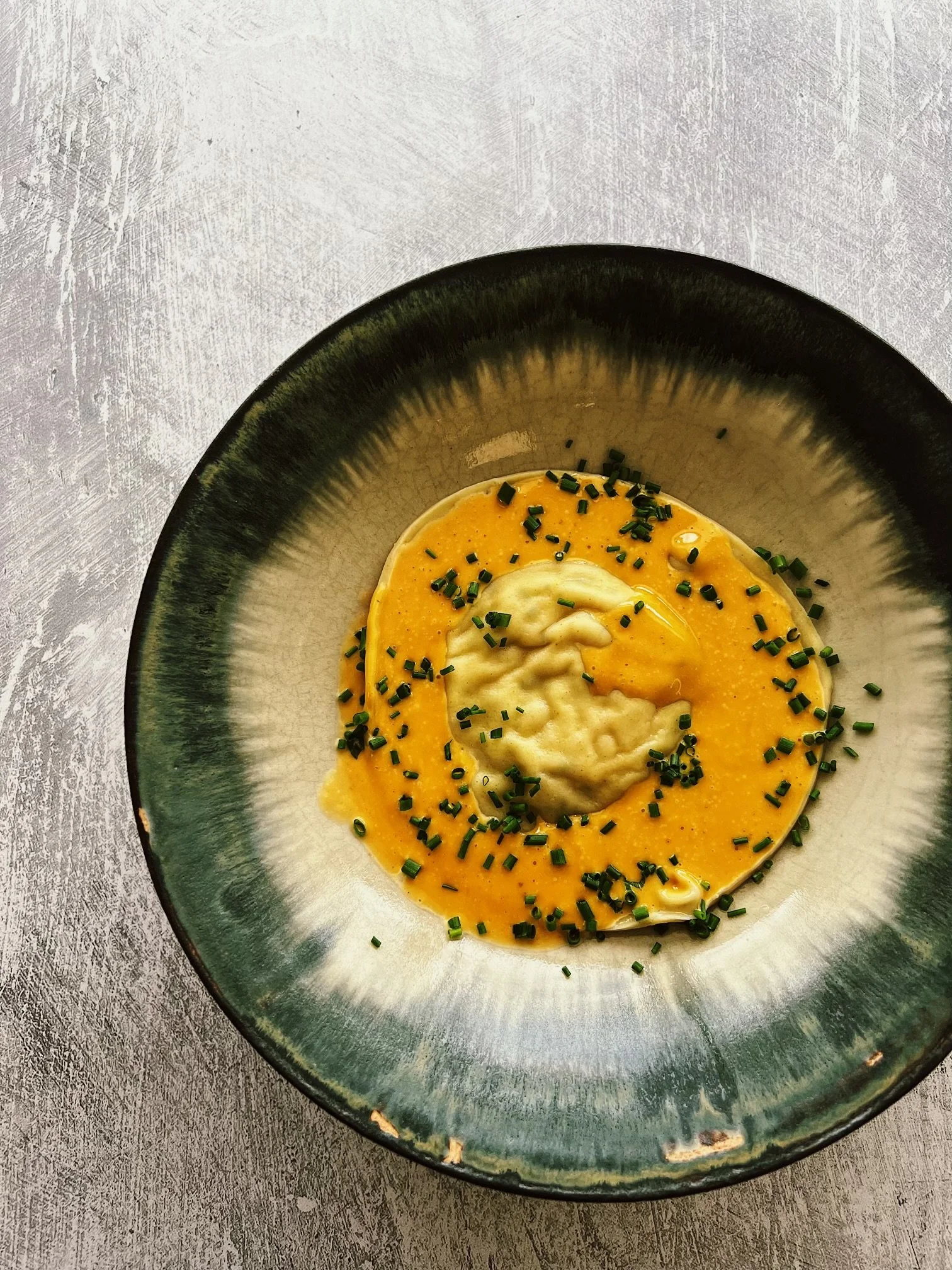 A dish of creamy mashed potatoes topped with melted cheese sauce, garnished with chopped chives, served in a black-rimmed bowl on a gray textured surface.