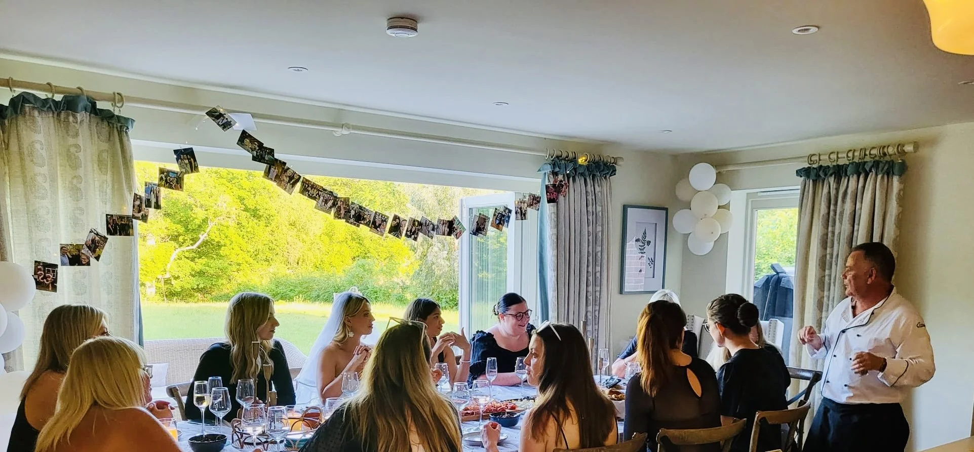 Indoor gathering of women celebrating, wedding reception or party, with chef speaking, decorated with hanging photos, windows showing green outdoor scenery, and balloons.