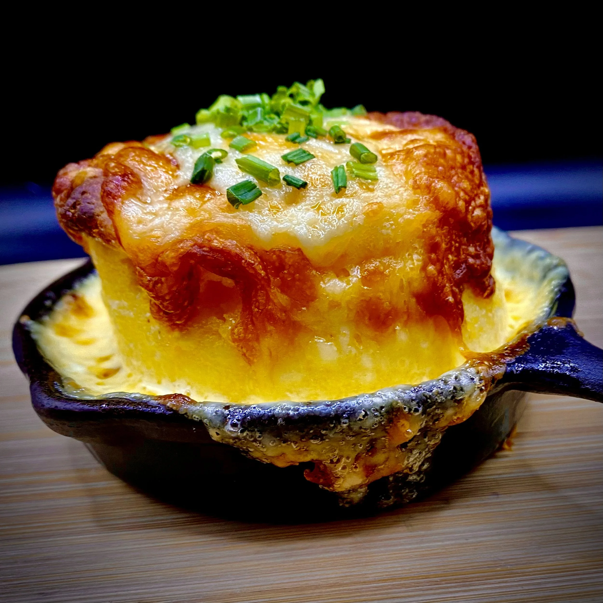 Close-up of a cheesy baked dish topped with chopped green onions, served in a black skillet on a wooden surface.