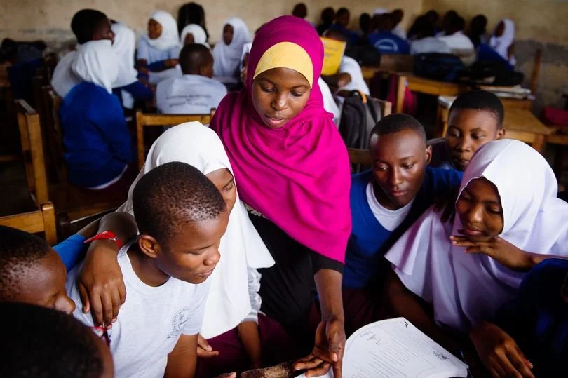A female teacher with a pink headscarf is helping a group of students in a classroom. The students, some wearing uniforms, are gathered around her and looking at a notebook or book. There are more students in the background sitting at desks.