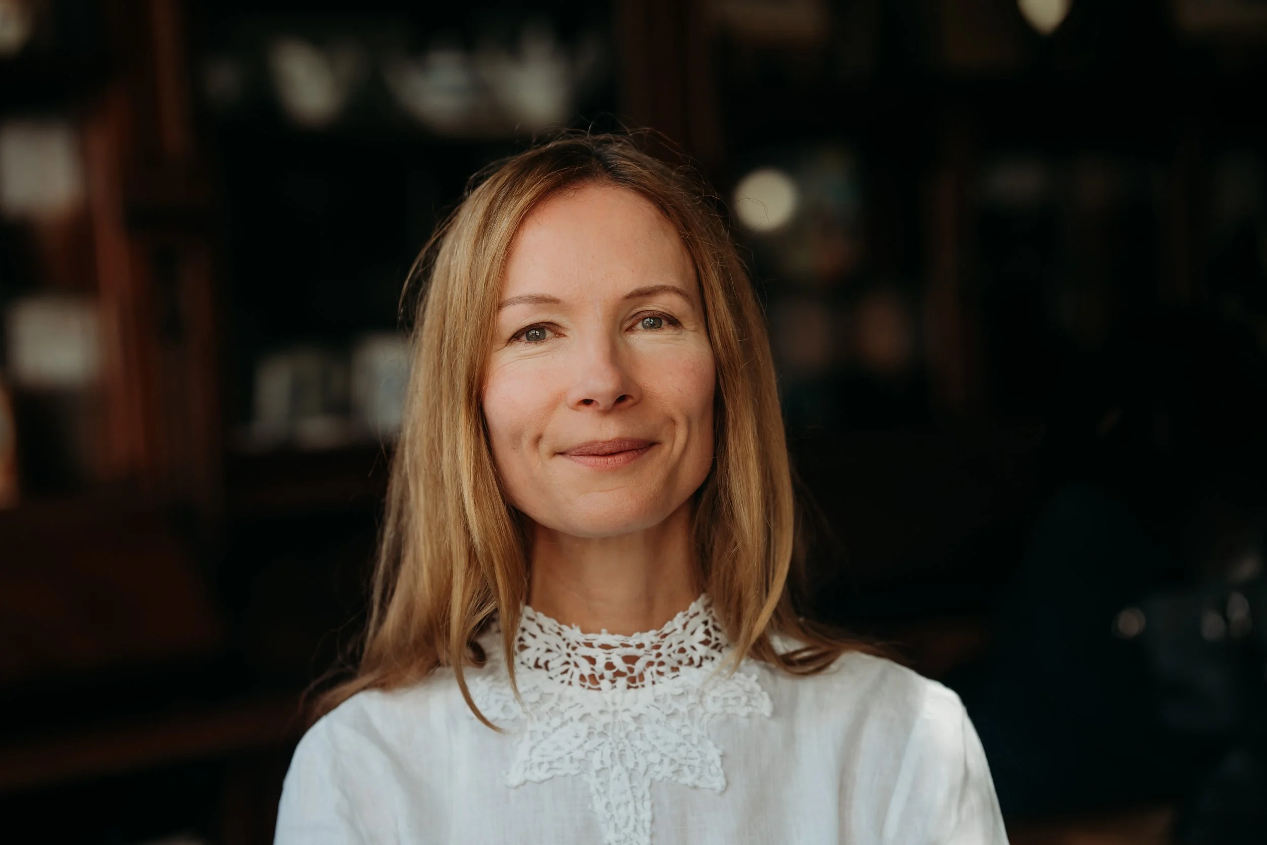 A woman with long, light brown hair and fair skin, wearing a white top with lace detailing at the neckline, smiling softly at the camera. The background is dark and blurred.