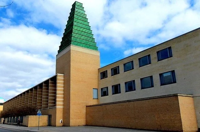 Modern brick building with a tall, pyramid-shaped, green glass tower against a partly cloudy sky.