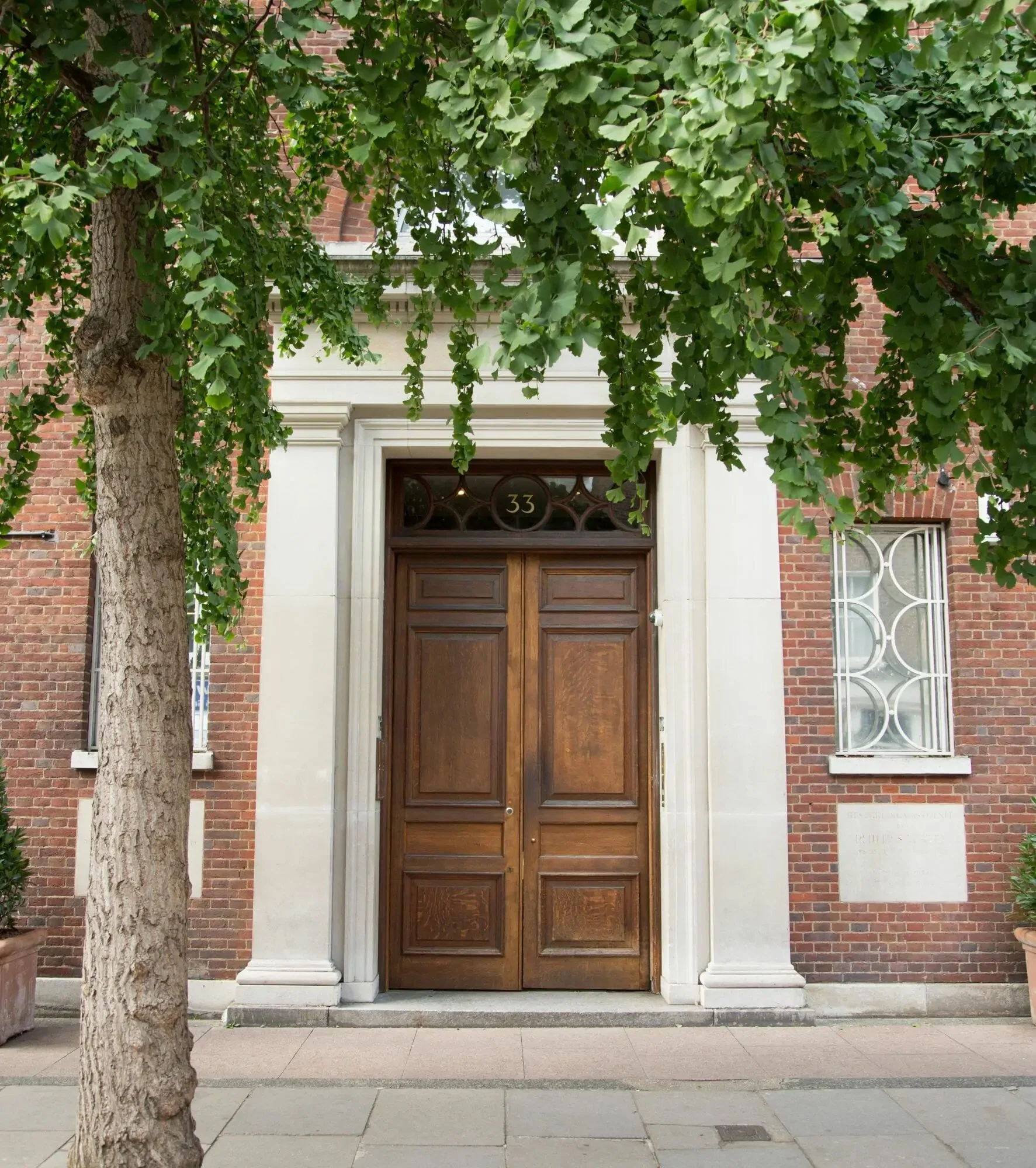 A brown wooden door with the number 33 above it, set in a building with brick walls and white stone framing. Lush green leafy tree branches hang in front, partially covering the door. The sidewalk is made of large gray tiles.