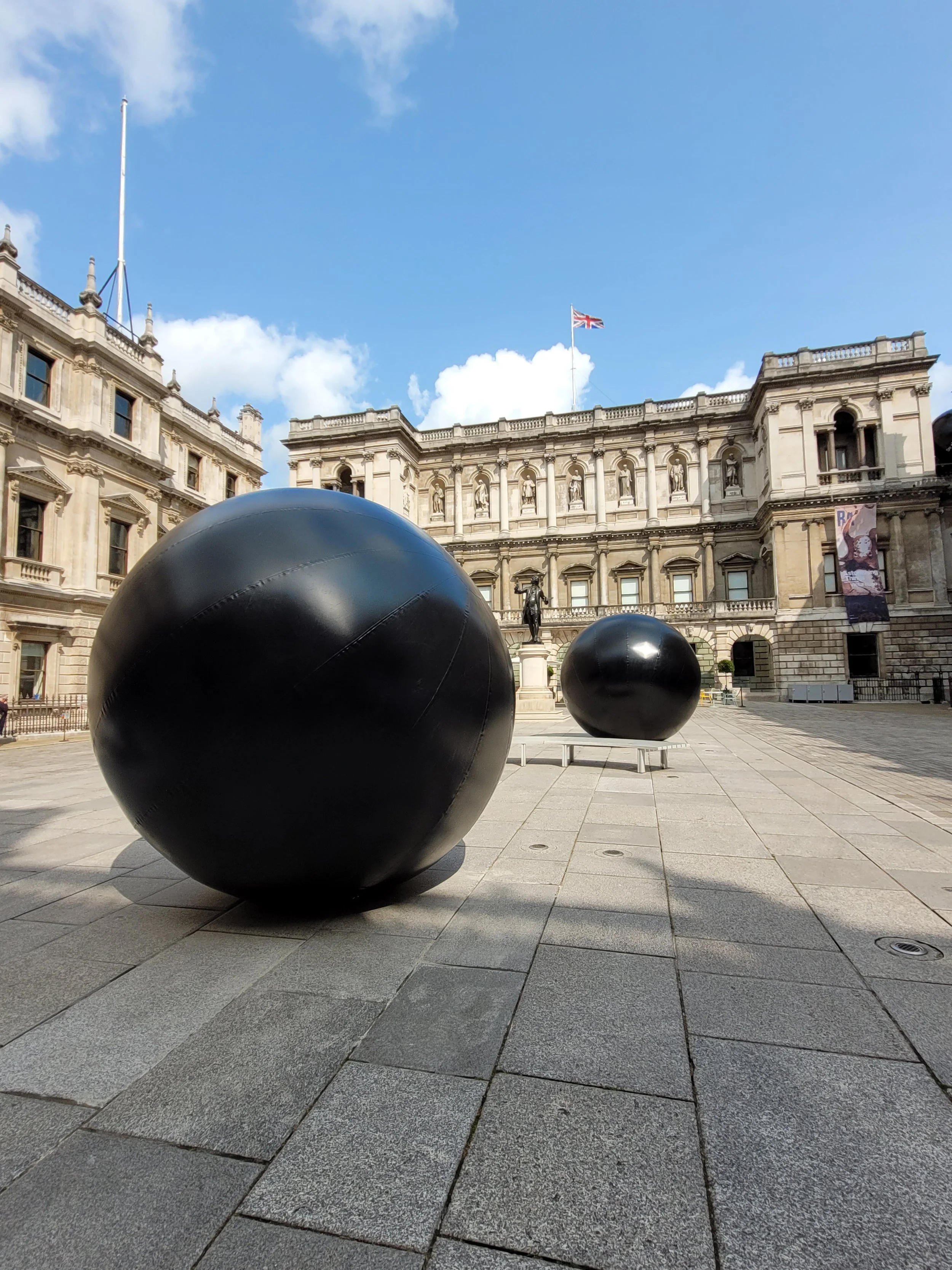 Large black spherical sculptures in a city square with historic buildings in the background under a blue sky.