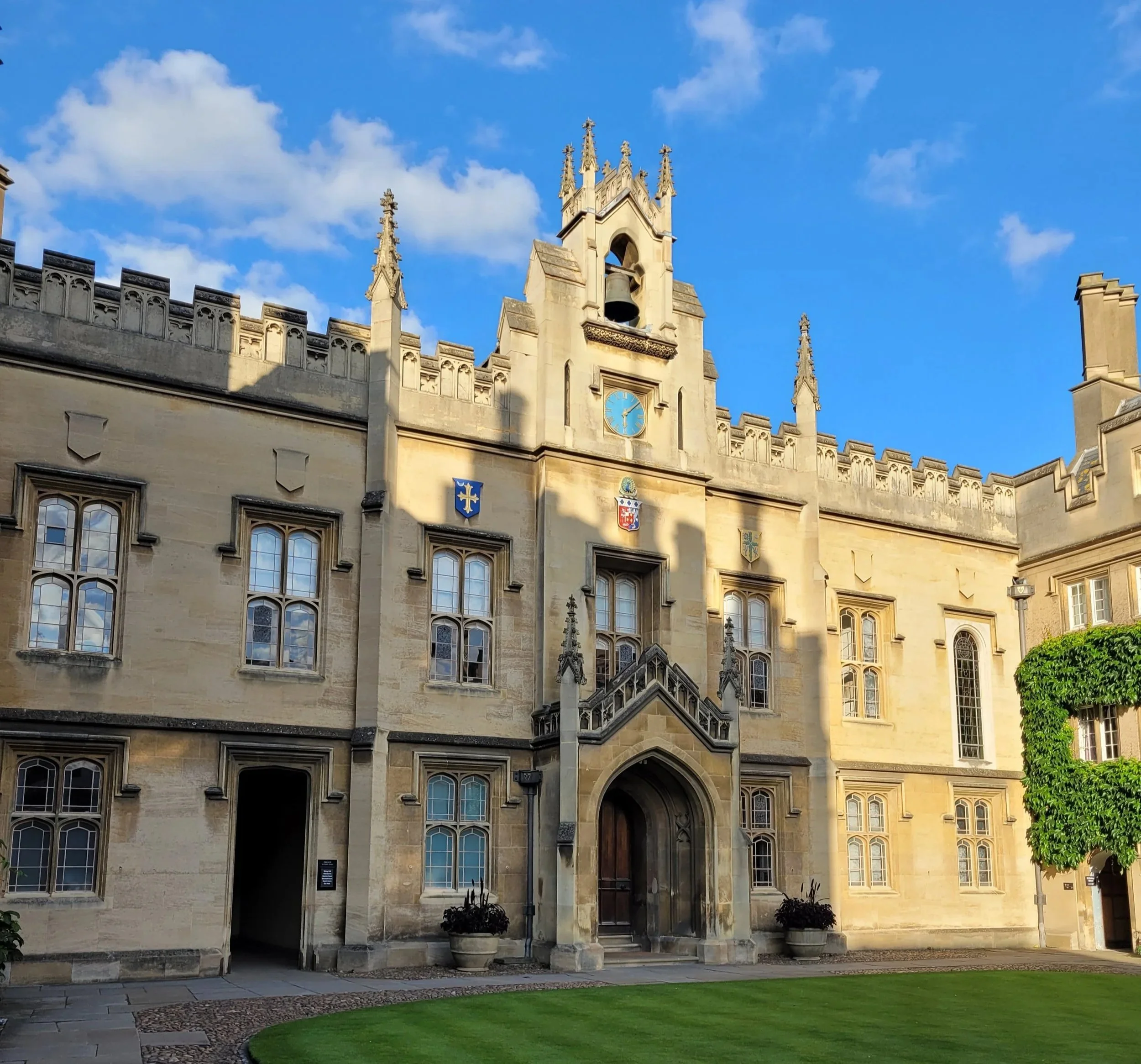 The image features the upper portion of a historic stone building with Gothic architectural details, including pointed arches, ornate spires, and crest shields on the facade. The building has large arched windows and a clock showing the time as approximately 11:10. The sky above is mostly blue with some fluffy white clouds.