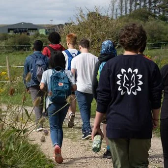 A group of young people walking outdoors on a dirt trail, carrying backpacks, surrounded by greenery.