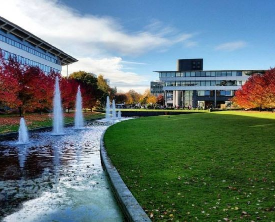 Modern office buildings with glass facades, a landscaped park with green grass, colorful autumn trees, and a water feature with fountains under a partly cloudy sky.
