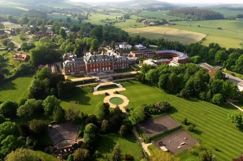 A large, historic mansion surrounded by well-maintained gardens and green lawns, with a circular driveway and multiple trees, set in a rural area with fields and forests in the background.