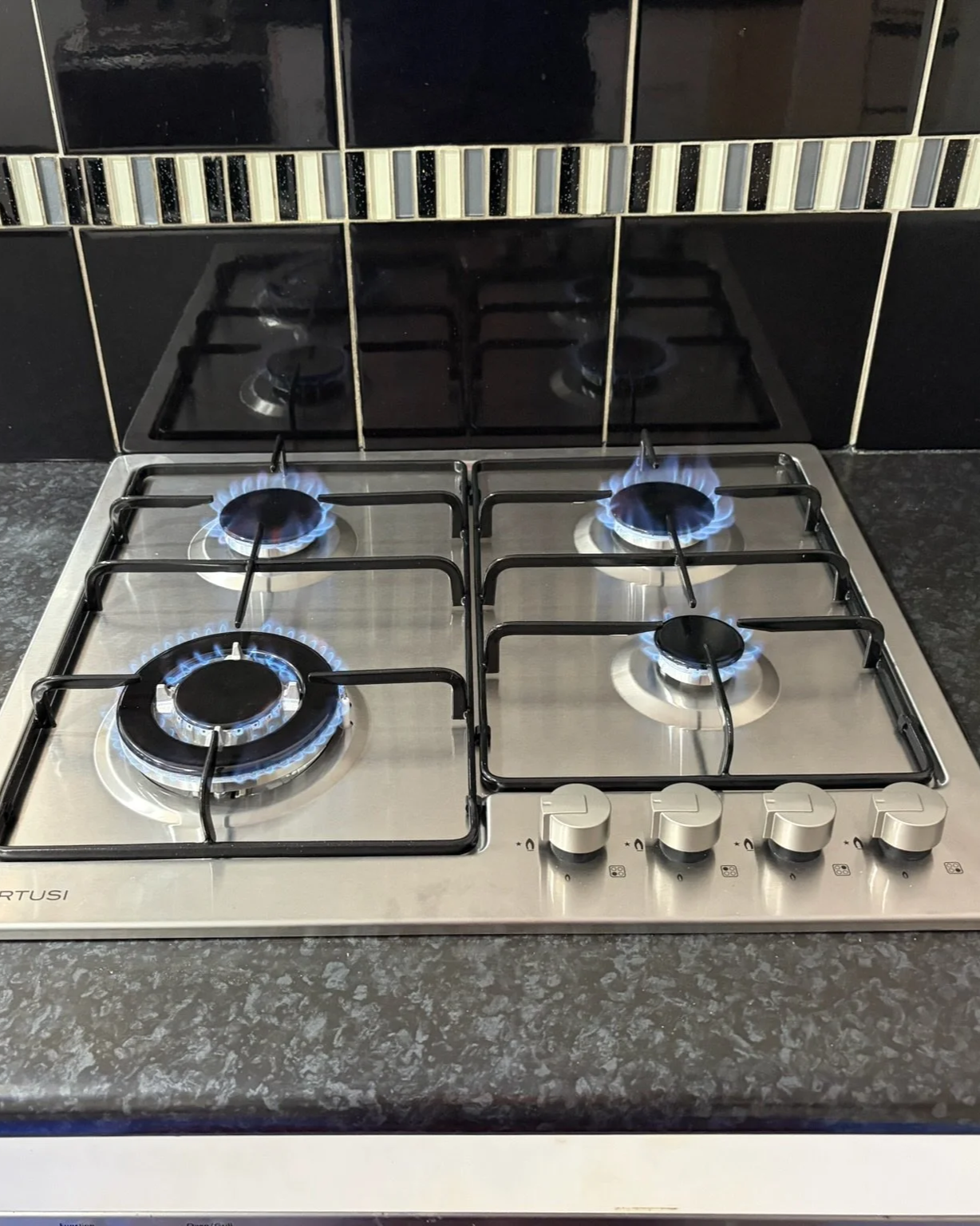 A stainless steel gas cooktop with four lit burners on a black granite countertop, with a black tiled backsplash featuring a decorative strip of small rectangular tiles.