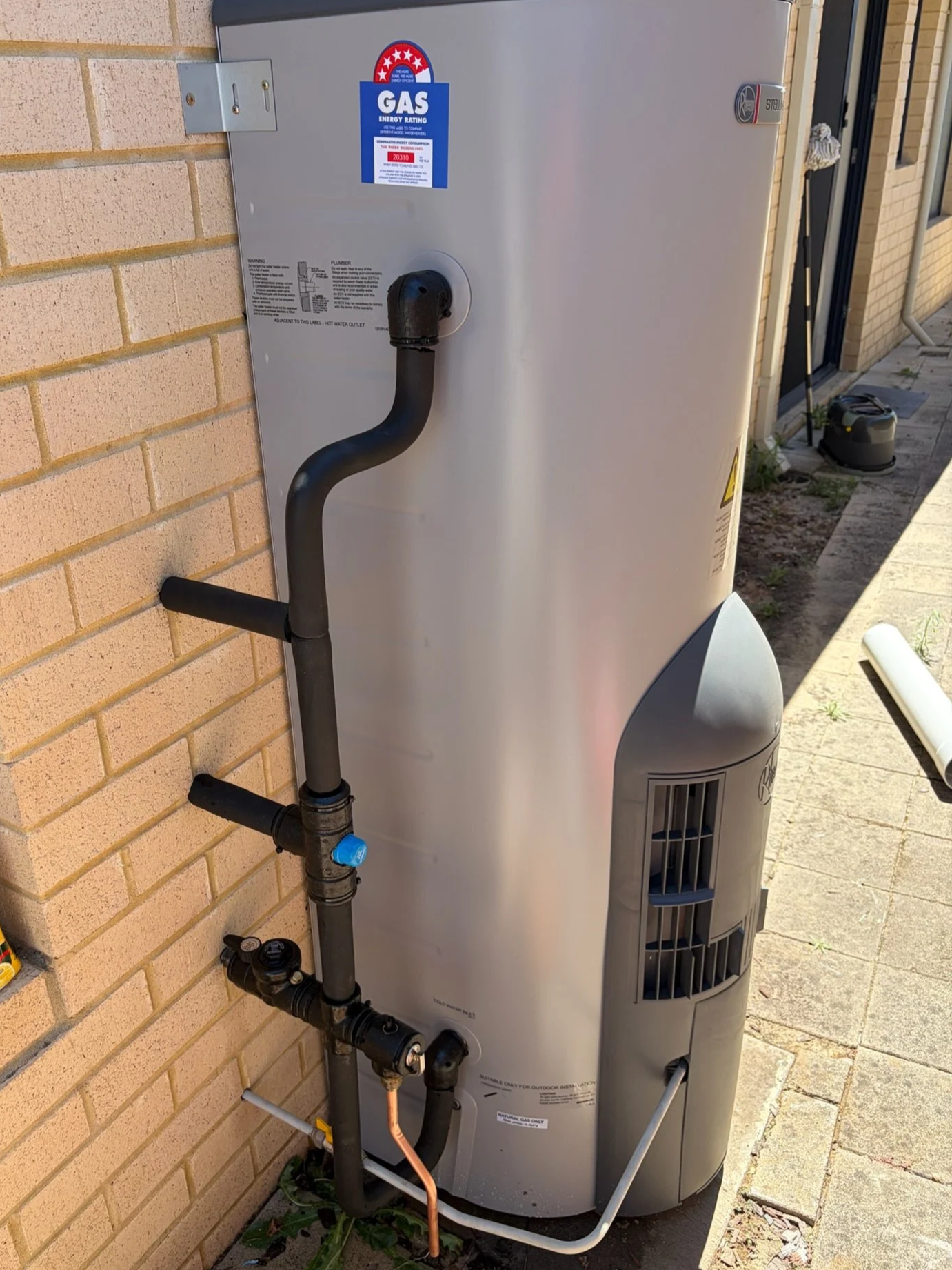An outdoor gas hot water heater with pipes connected to it, installed near a brick wall and beside a sidewalk.