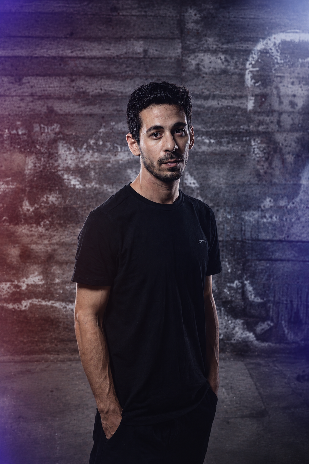 A young man with dark, curly hair and a goatee, wearing a black t-shirt, standing against a textured concrete wall with colored lighting effects.