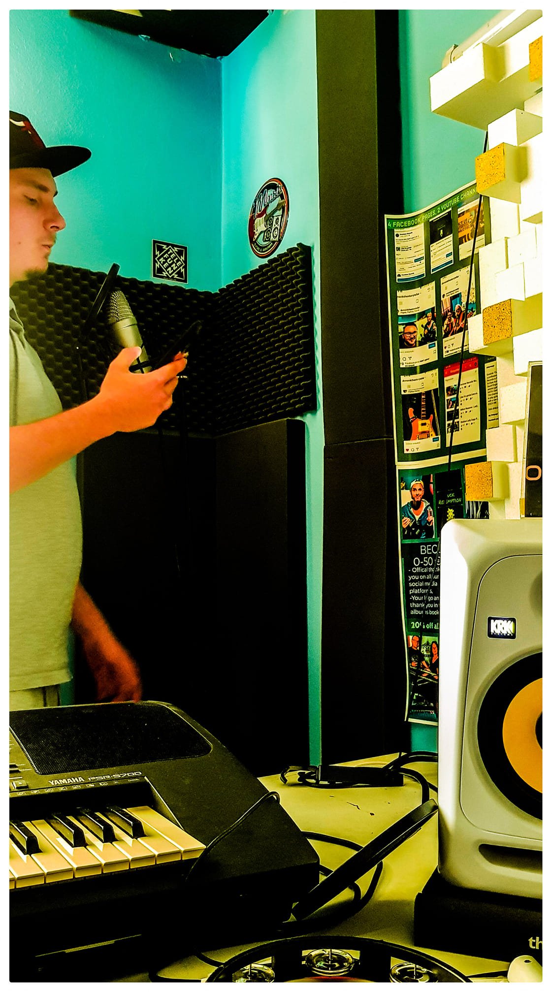 Person standing in a music studio, holding a phone, with a microphone, speaker, and DJ equipment around.