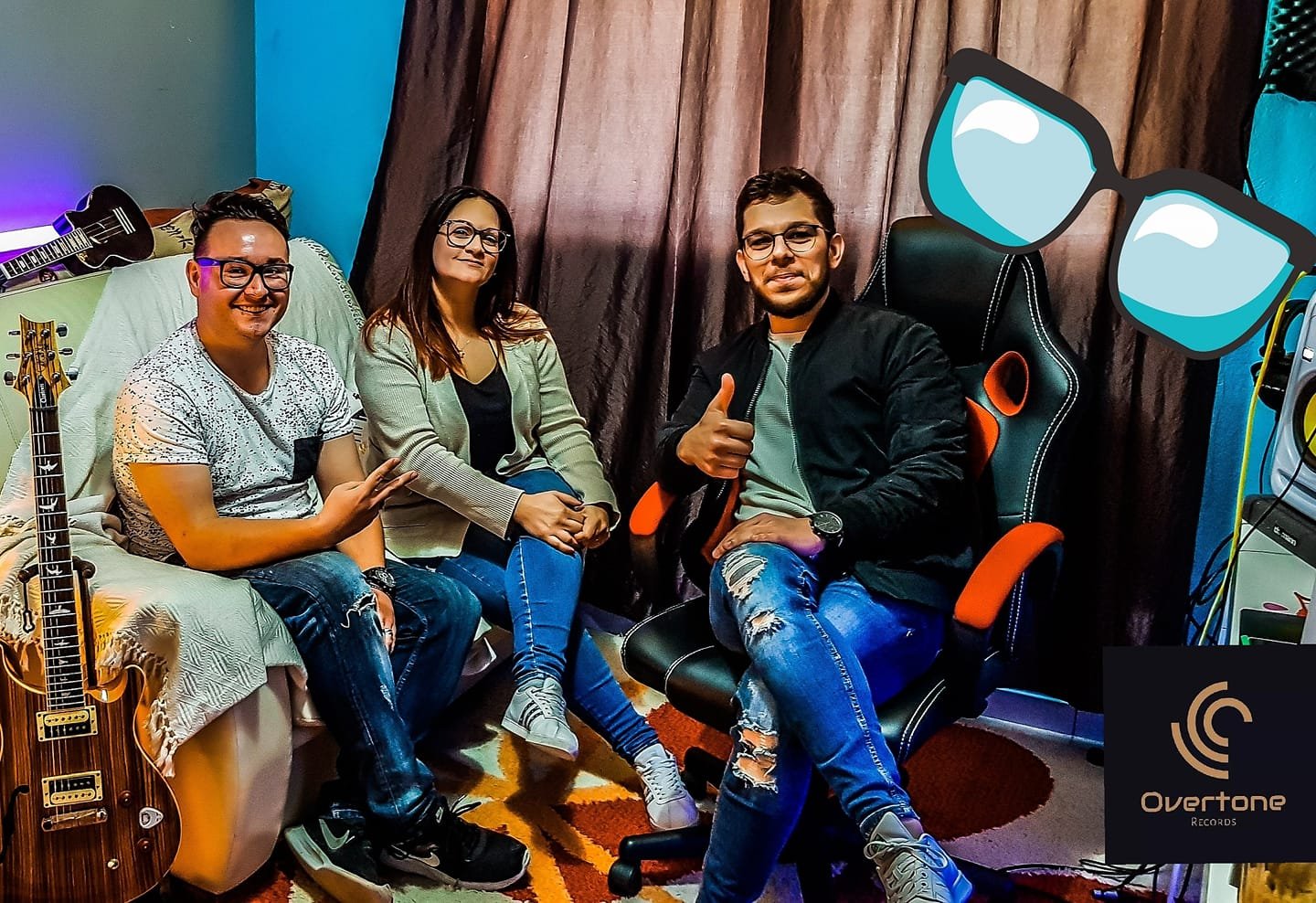 Four people sitting in a music studio with guitars and music equipment, one person giving a thumbs-up while another makes a peace sign.