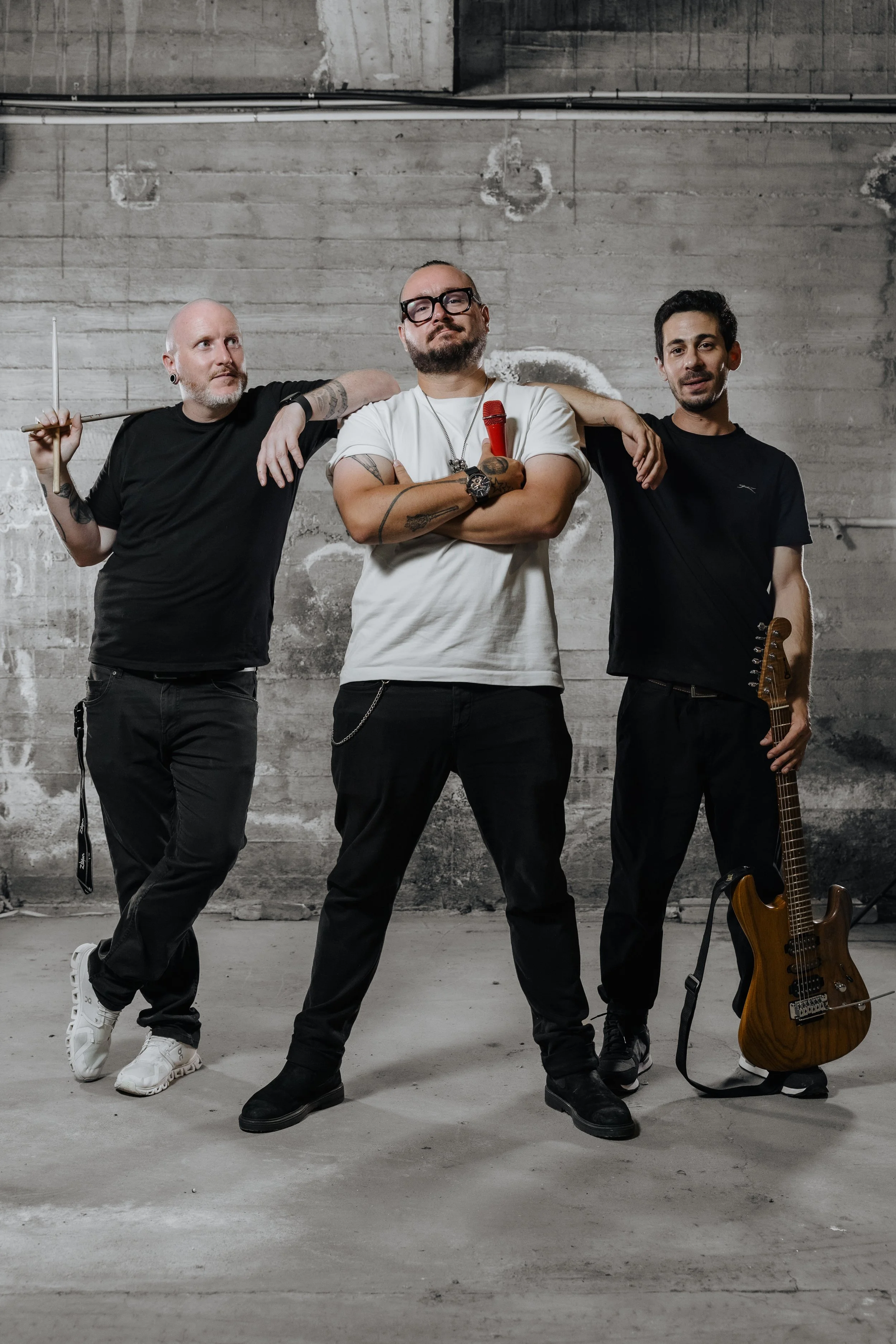 Three men standing in front of a concrete wall. The man in the center has his arms crossed and is holding a red microphone. The man on the left is holding a drumstick, and the man on the right is holding an electric bass guitar. All three appear rela