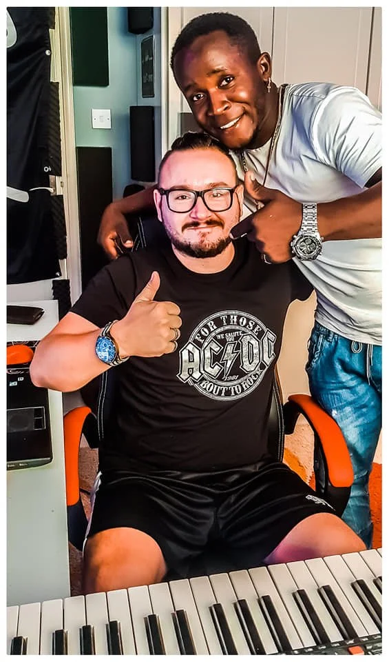 Two men posing together indoors. One sitting at a keyboard, giving a thumbs-up, wearing glasses and a black AC/DC T-shirt, with a blue watch. The other man standing behind him, smiling, with arm around the seated man, wearing a white T-shirt, jewelry