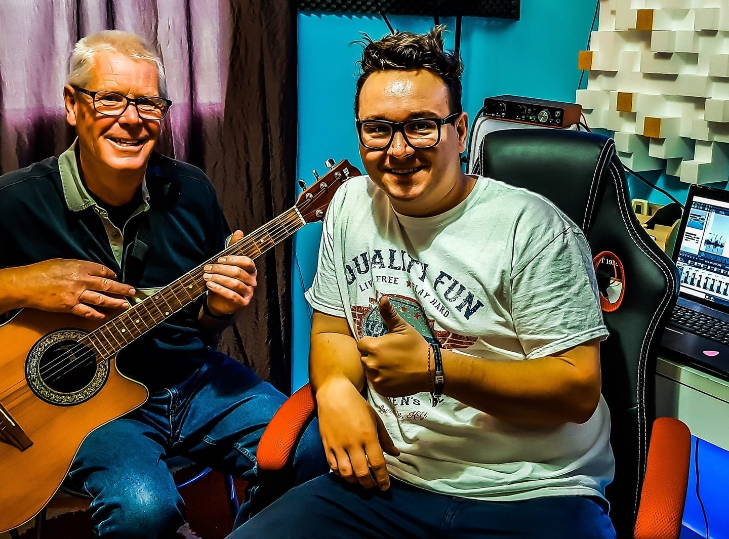 Two men in a recording studio, one playing an acoustic guitar and the other sitting in a chair giving a thumbs-up, smiling.