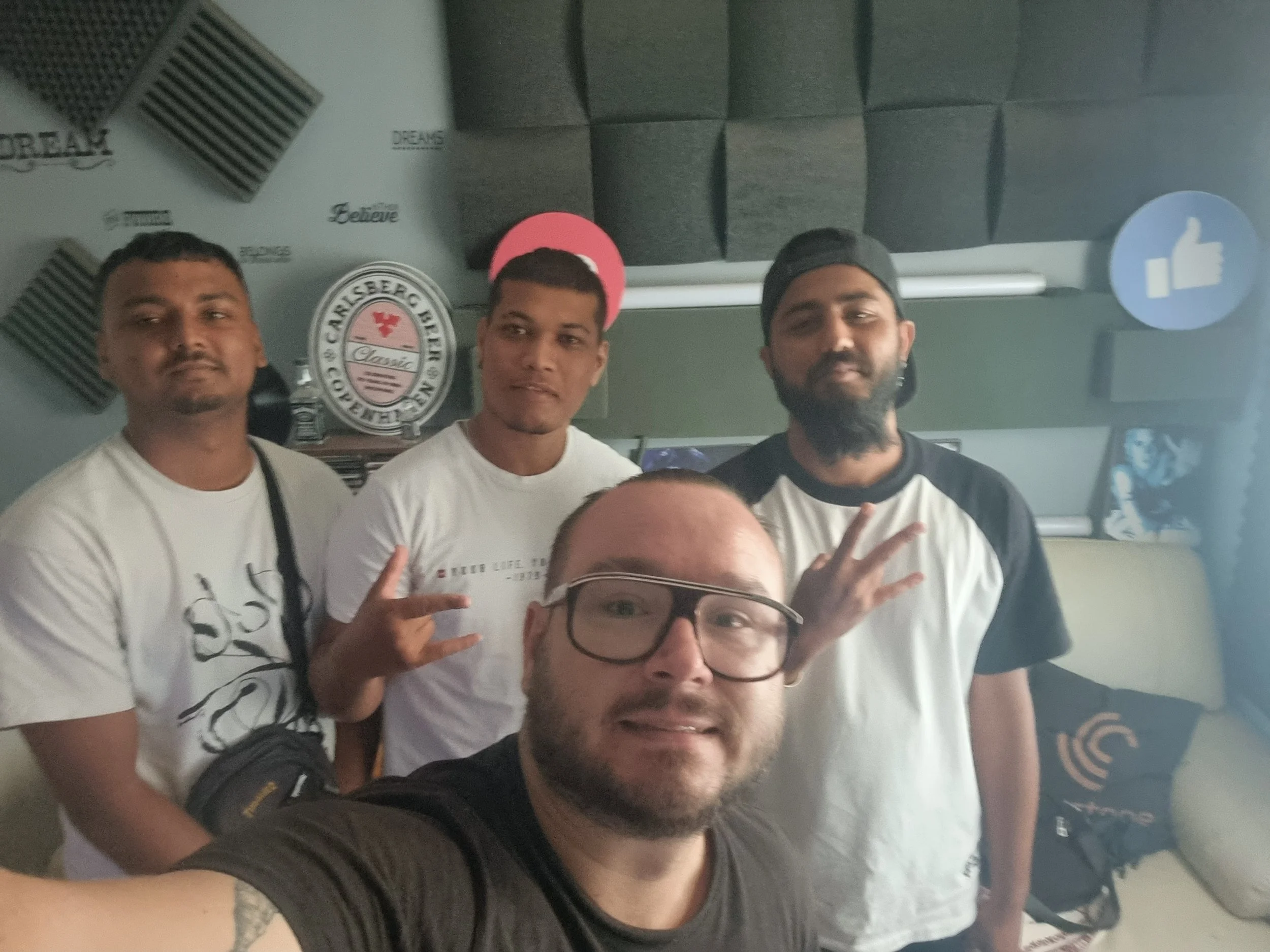 Group of five men taking a selfie in a studio with soundproof felt panels, a wall with logos, and icons including a thumbs-up sign.