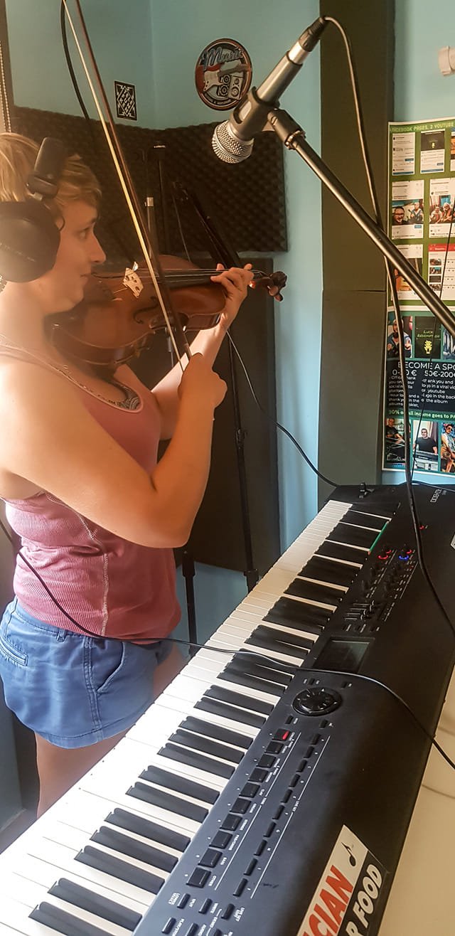 A woman wearing headphones playing a violin in a recording studio with a keyboard in front of her and a microphone overhead.