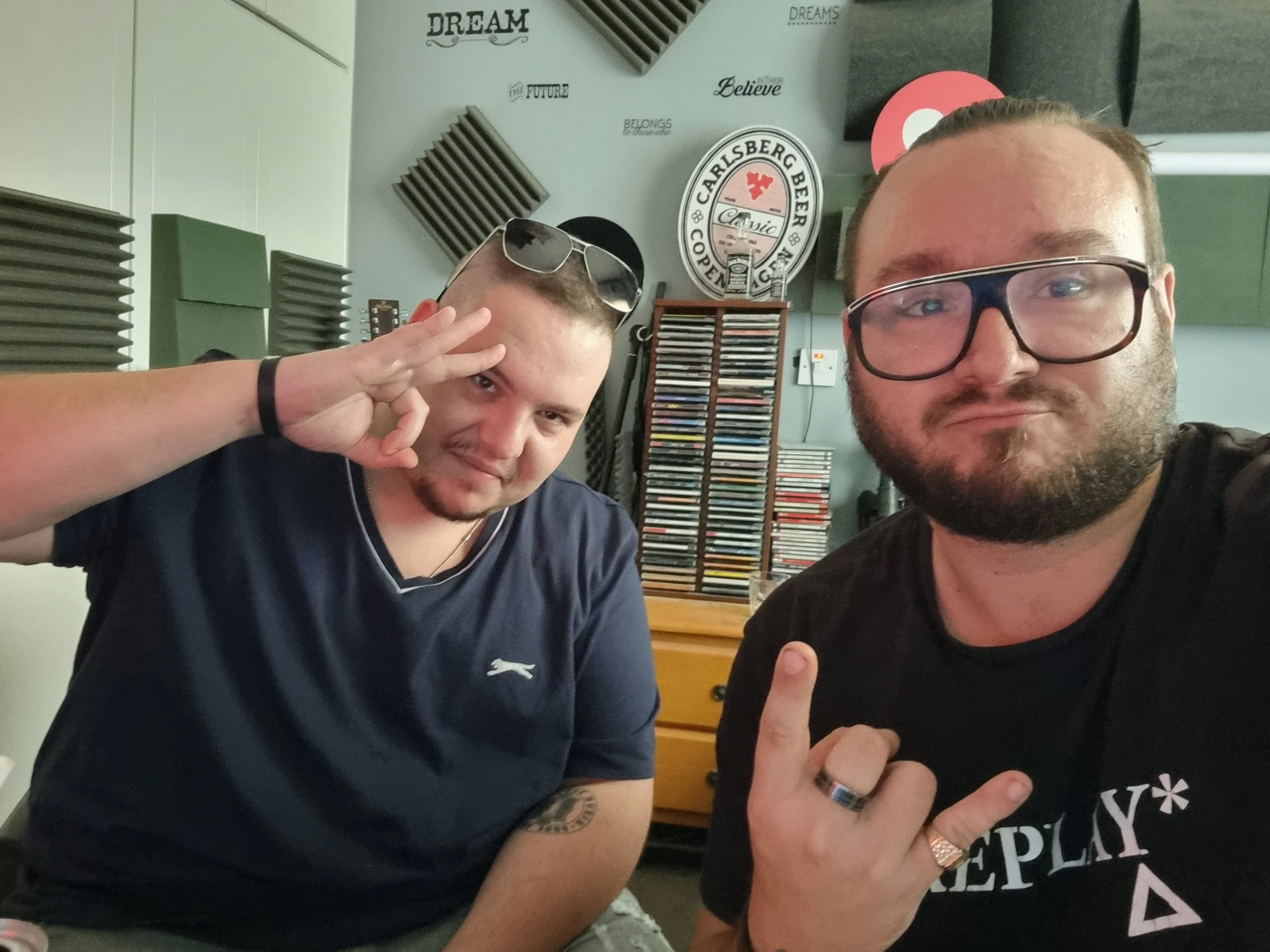 Two young men taking a selfie in a room with gray walls decorated with foam soundproof panels, wall decals, and shelves filled with CDs. One man is wearing glasses and a black T-shirt, while the other is making a peace sign. They appear to be in a mu