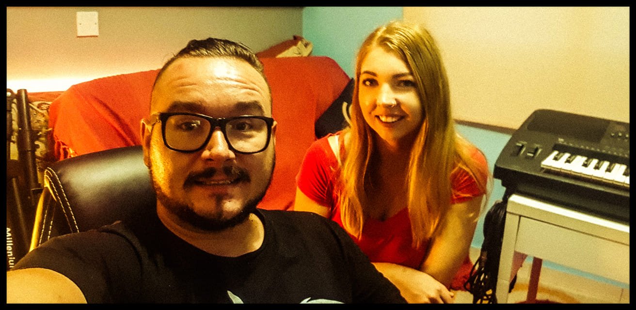 A man with glasses and a beard taking a selfie with a woman with long blonde hair in a room with a keyboard on a stand and a red sofa in the background.