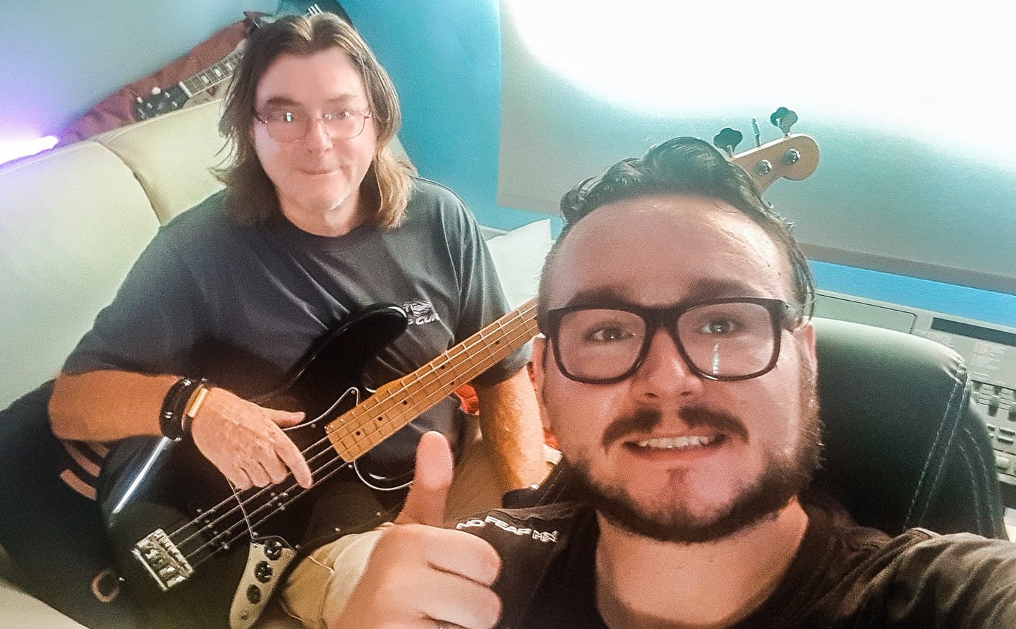 Two men smiling, one with dark hair and glasses giving a thumbs up, the other with long light hair and glasses holding a bass guitar, in a music studio with a computer, sound equipment, and a guitar in the background.