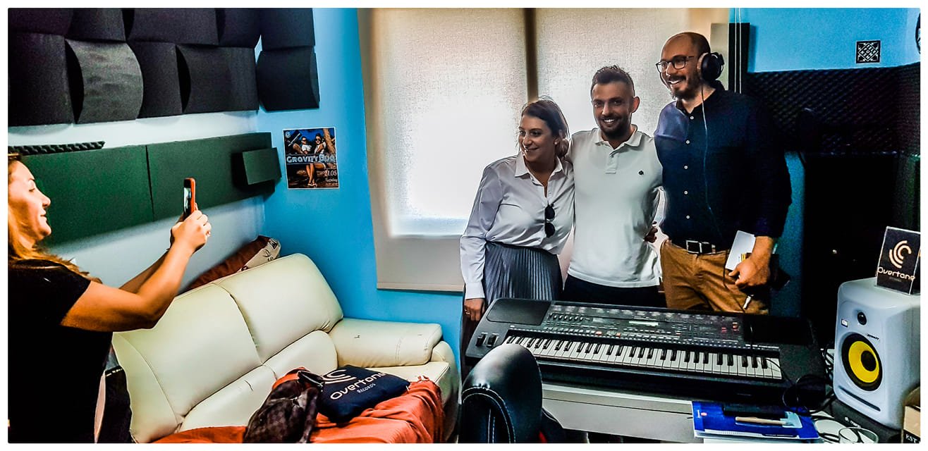 Three people standing close together in a music studio, posing for a photo, with a woman taking the picture. The studio has a keyboard, speakers, and soundproof foam on the walls.