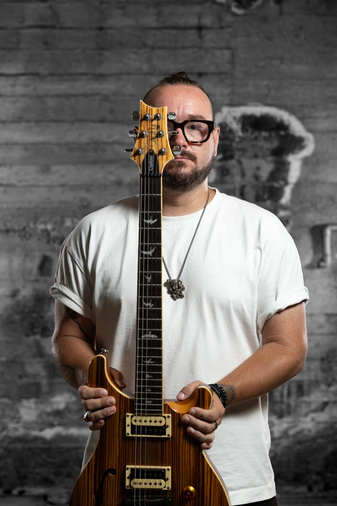 A man with glasses and a beard holding an electric guitar standing in front of a textured gray wall with a blurred portrait of a person's face in the background.