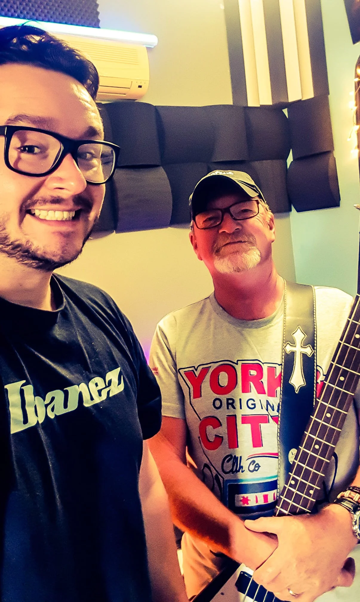 Two men smiling, one wearing glasses and a black shirt with white text, and the other wearing glasses, a cap, and a t-shirt with red and black text, holding a guitar in a music studio with soundproofing panels and a neon light in the background.