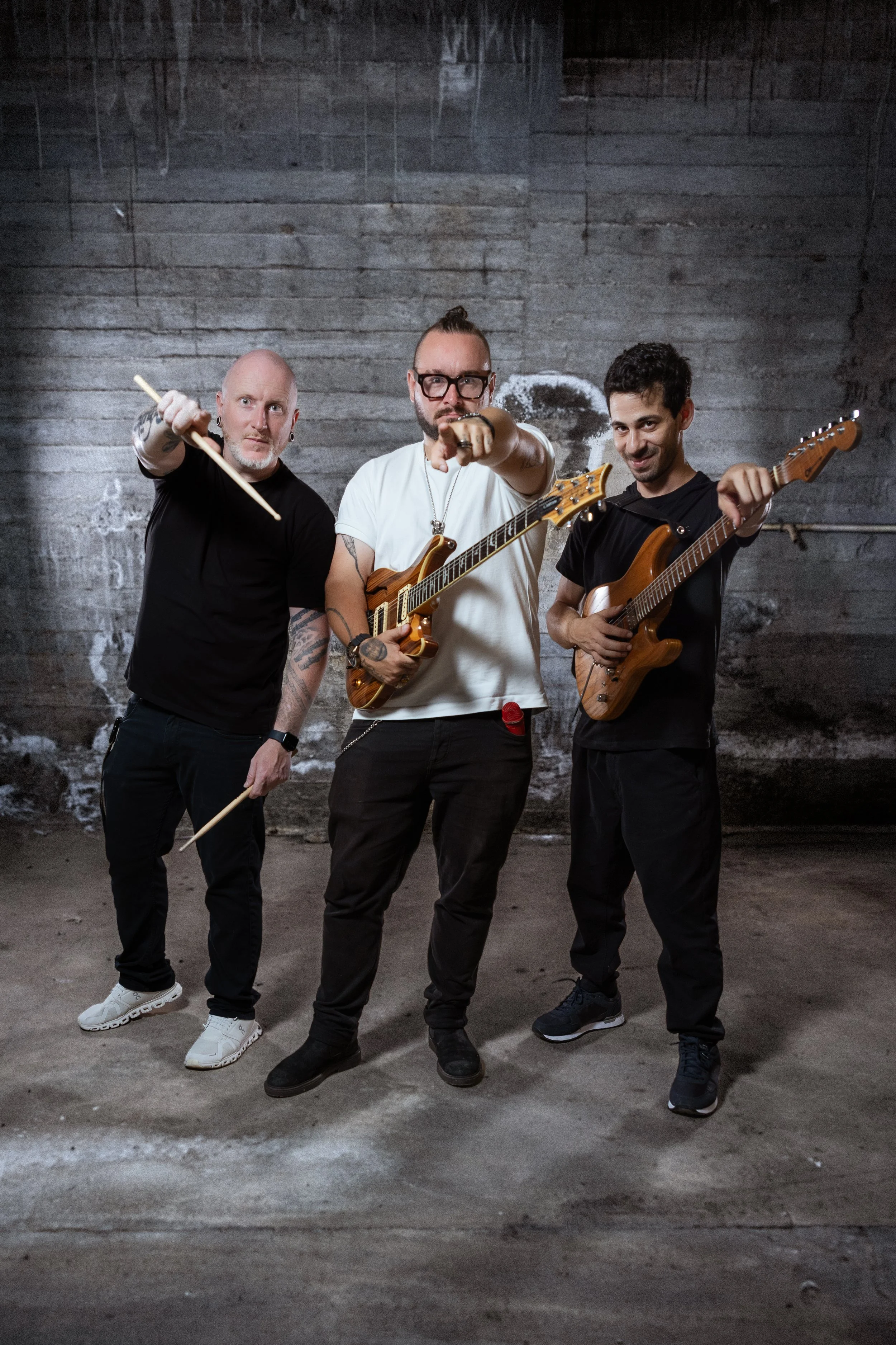 Three men in casual clothing posing with musical instruments in a gritty, industrial setting. The man on the left holds drumsticks, the middle man has an electric guitar, and the man on the right holds an electric guitar, all standing against a textu