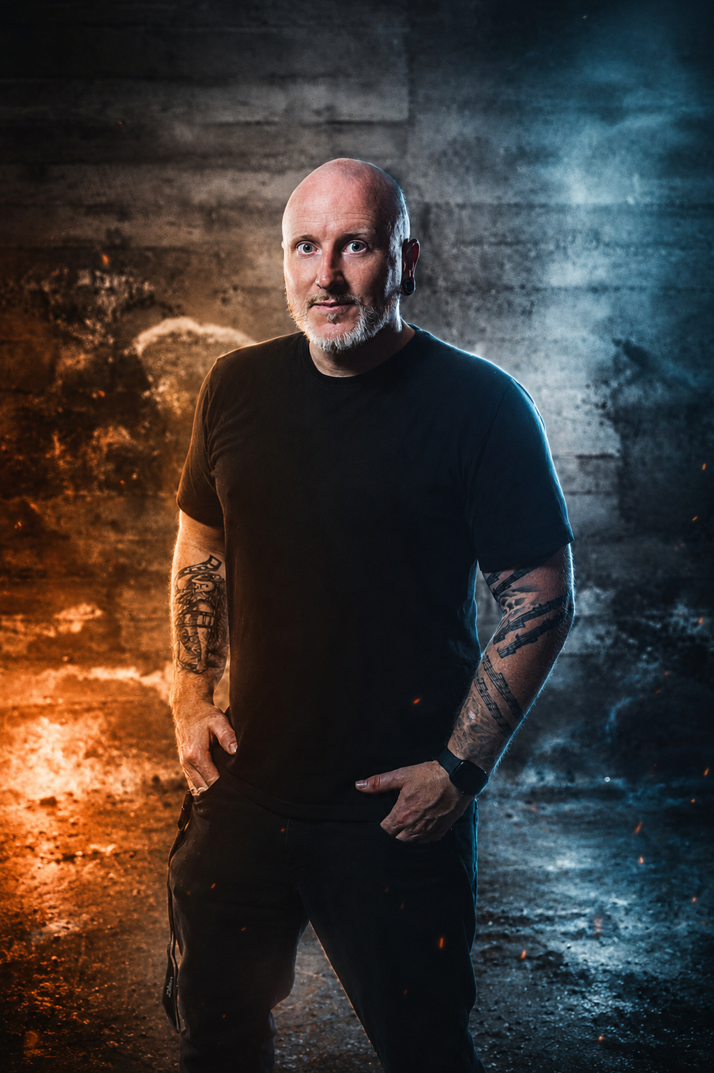 A man with a beard and tattoos on his arms wearing a black t-shirt and black pants, standing with hands in pockets against a dark background with orange and blue lighting effects.