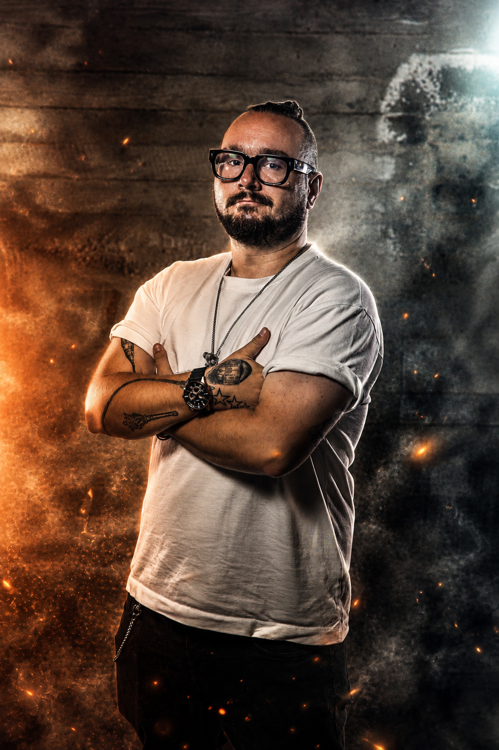 A man with glasses and a beard, wearing a white t-shirt with rolled-up sleeves, standing with crossed arms against a dark background with fiery effects and sparks.