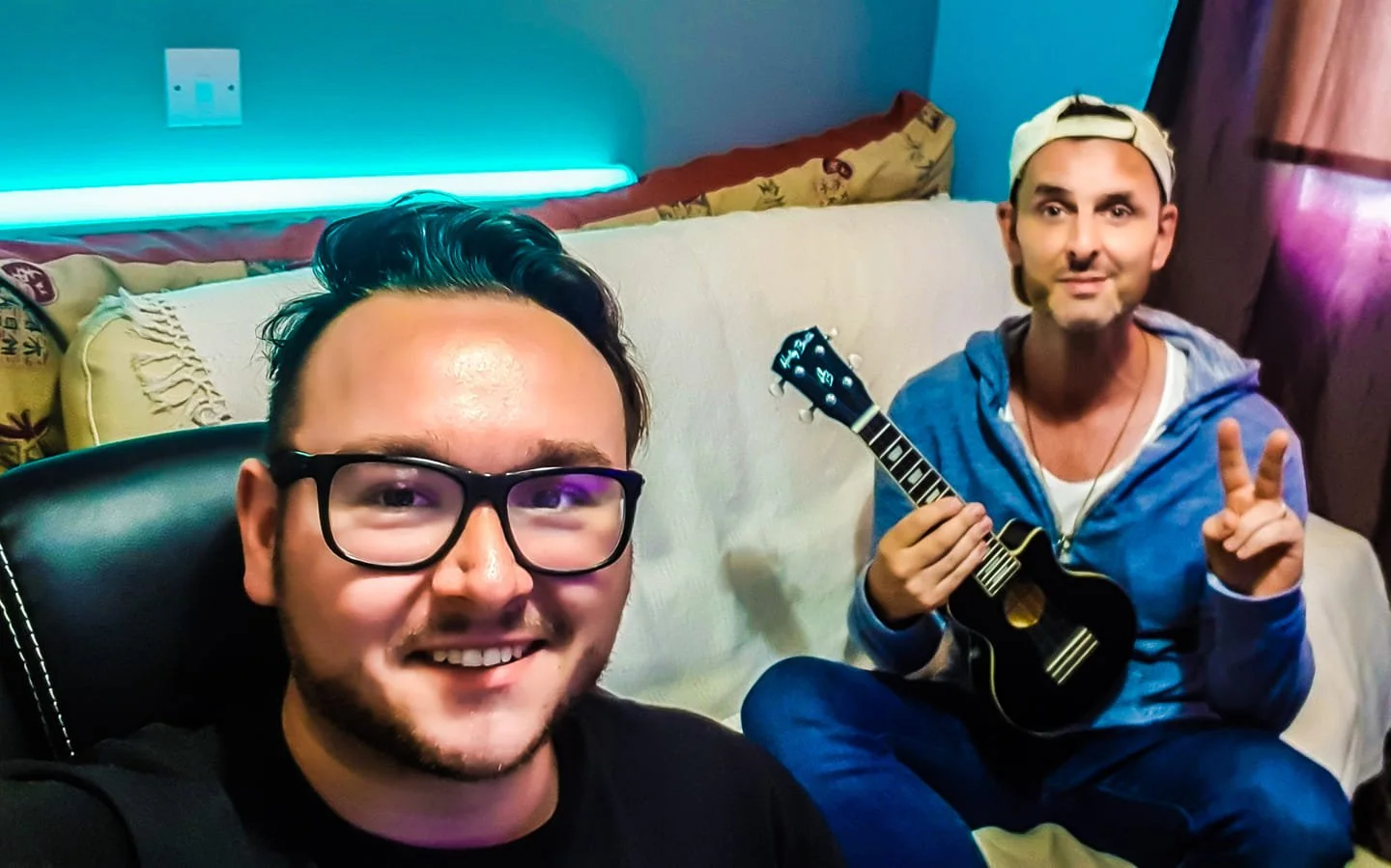 Two men sitting on a white sofa in a room with colorful lighting. The man in the foreground has glasses and dark hair, smiling at the camera. The man in the background is holding a small black ukulele, wearing a backwards cap, blue hoodie, and making