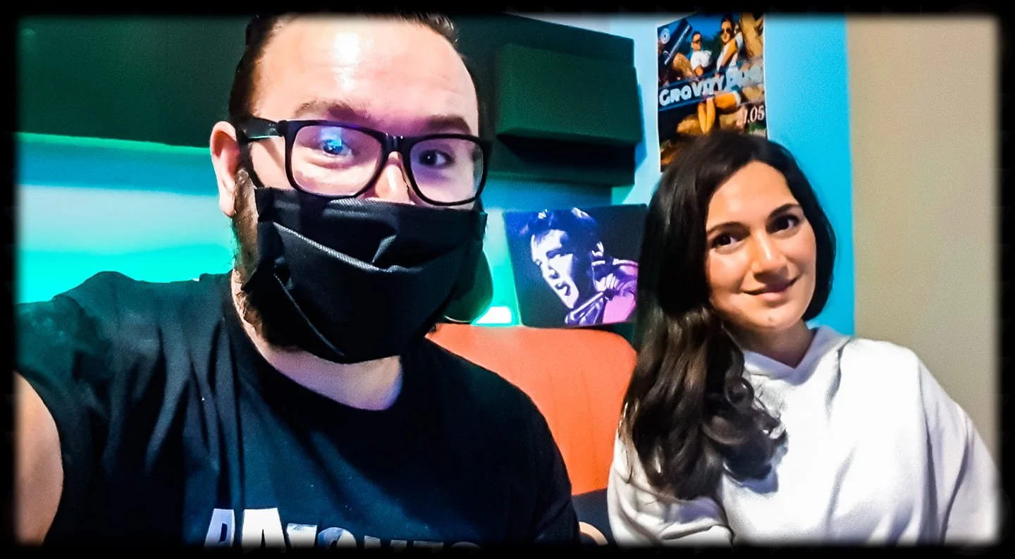 A man and a woman taking a selfie together indoors. The man is wearing glasses, a face mask, and a black T-shirt. The woman has long dark hair and is wearing a light-colored hoodie. Posters are visible on the wall behind them.