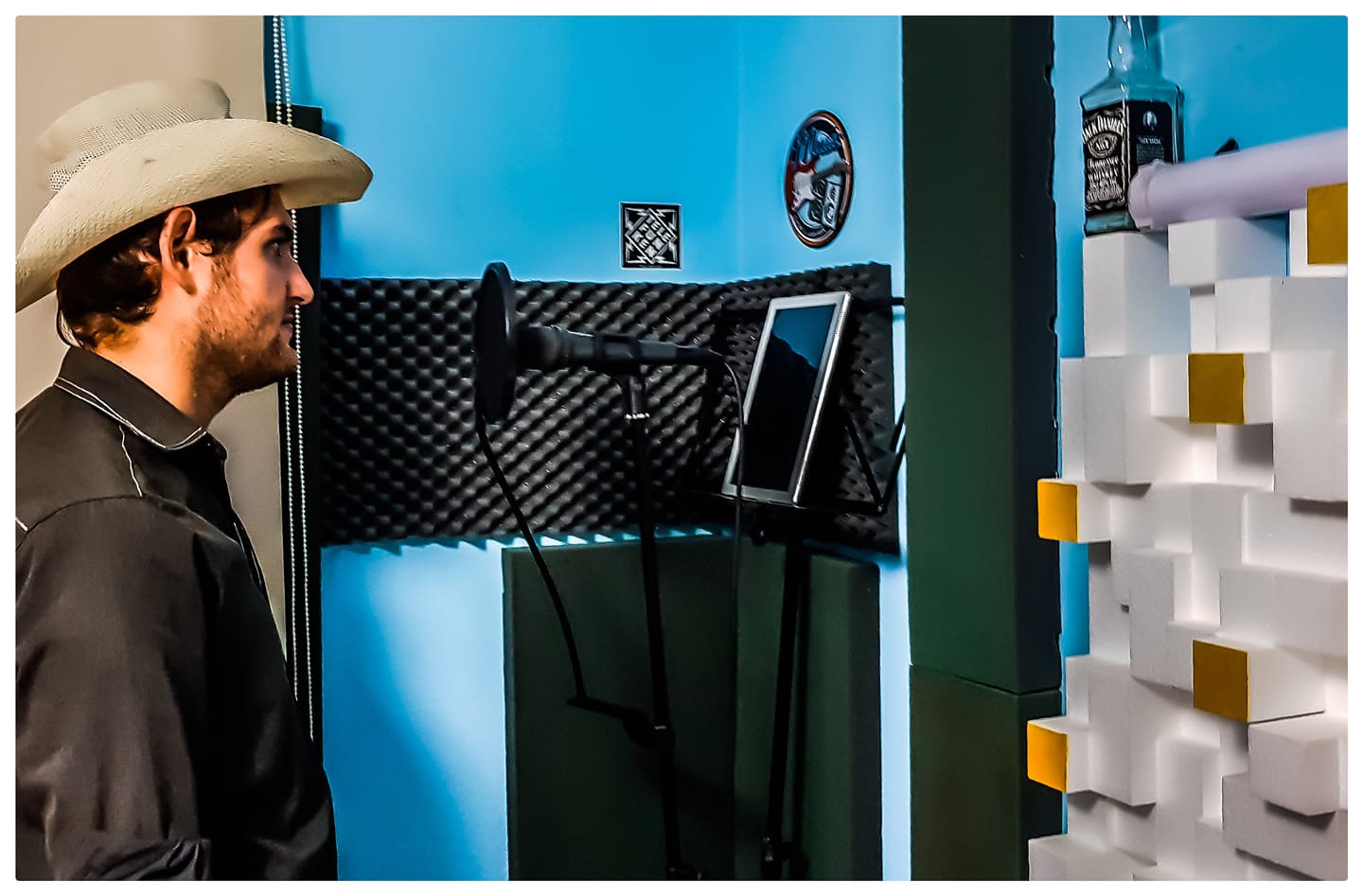 A man wearing a cowboy hat and a black jacket standing in a recording studio with a microphone, a small monitor, and acoustic foam panels on the walls.