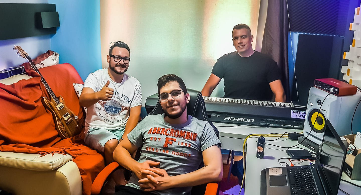 Four young men in a music studio, two sitting and one standing behind a keyboard, with musical equipment and a guitar in the background.