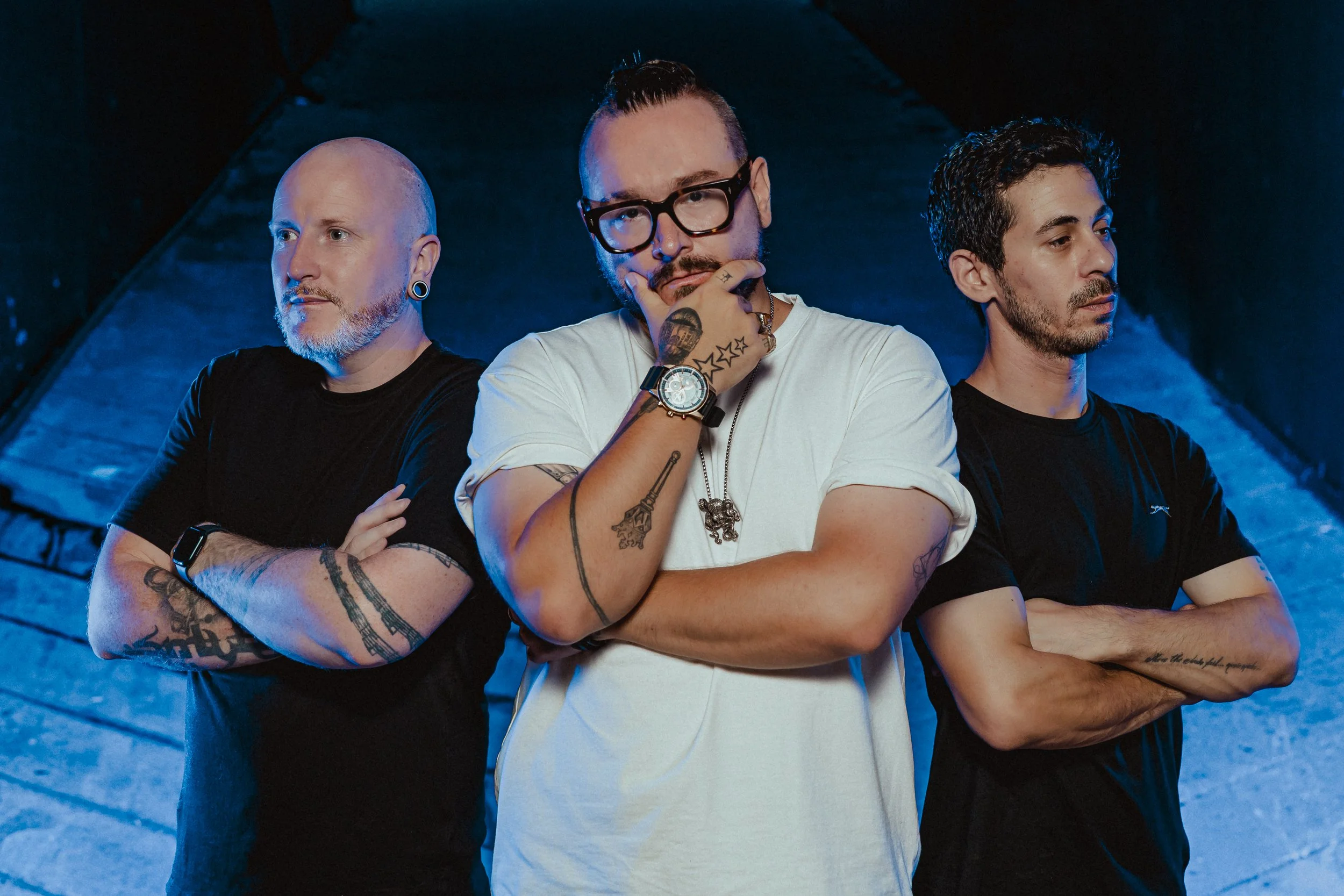 Three men stand with their arms crossed and one man in the middle holding his hand near his chin, in a dark setting with blue lighting.